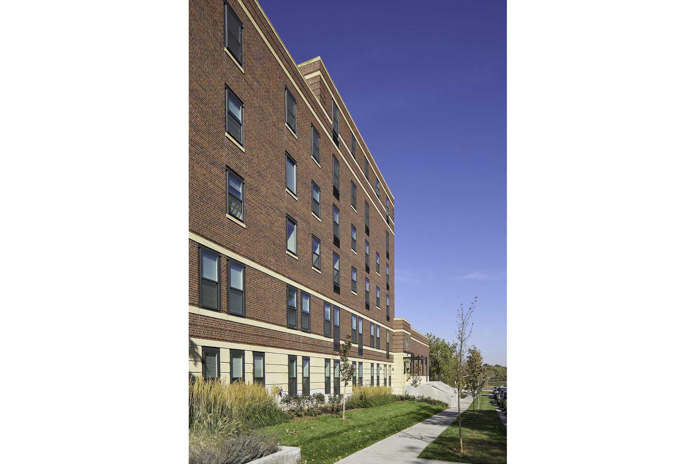 A three-story brick building with large windows and a landscaped walkway. The structure features a classic architectural design and is surrounded by greenery. The sky is clear and blue, enhancing the building's attractive appearance.