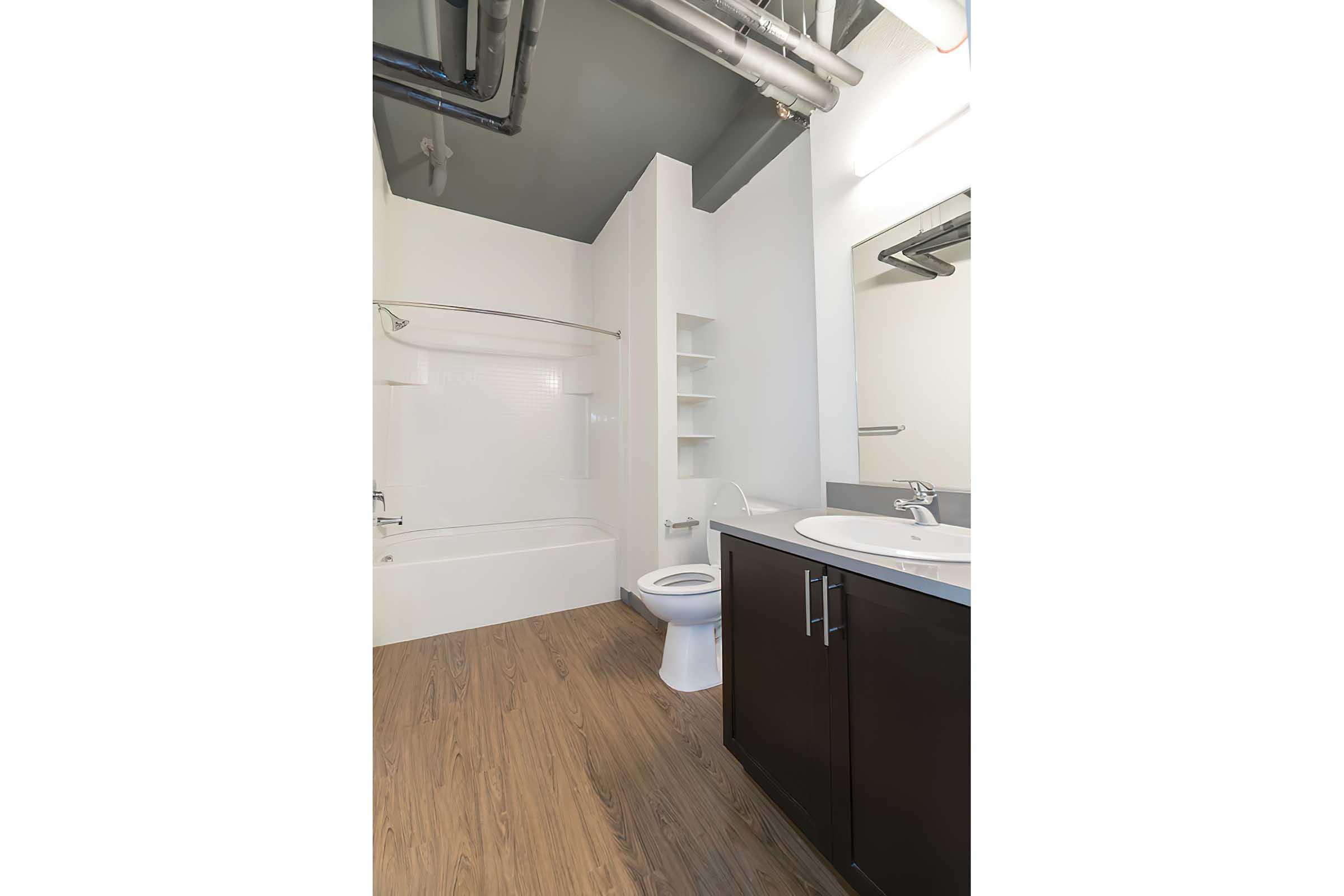 A modern bathroom featuring a bathtub with shower, a white toilet, a single vanity sink with a mirror above, and shelves on the wall. The space has light-colored walls and wooden flooring, complemented by exposed pipes on the ceiling, creating a clean and minimalist design.