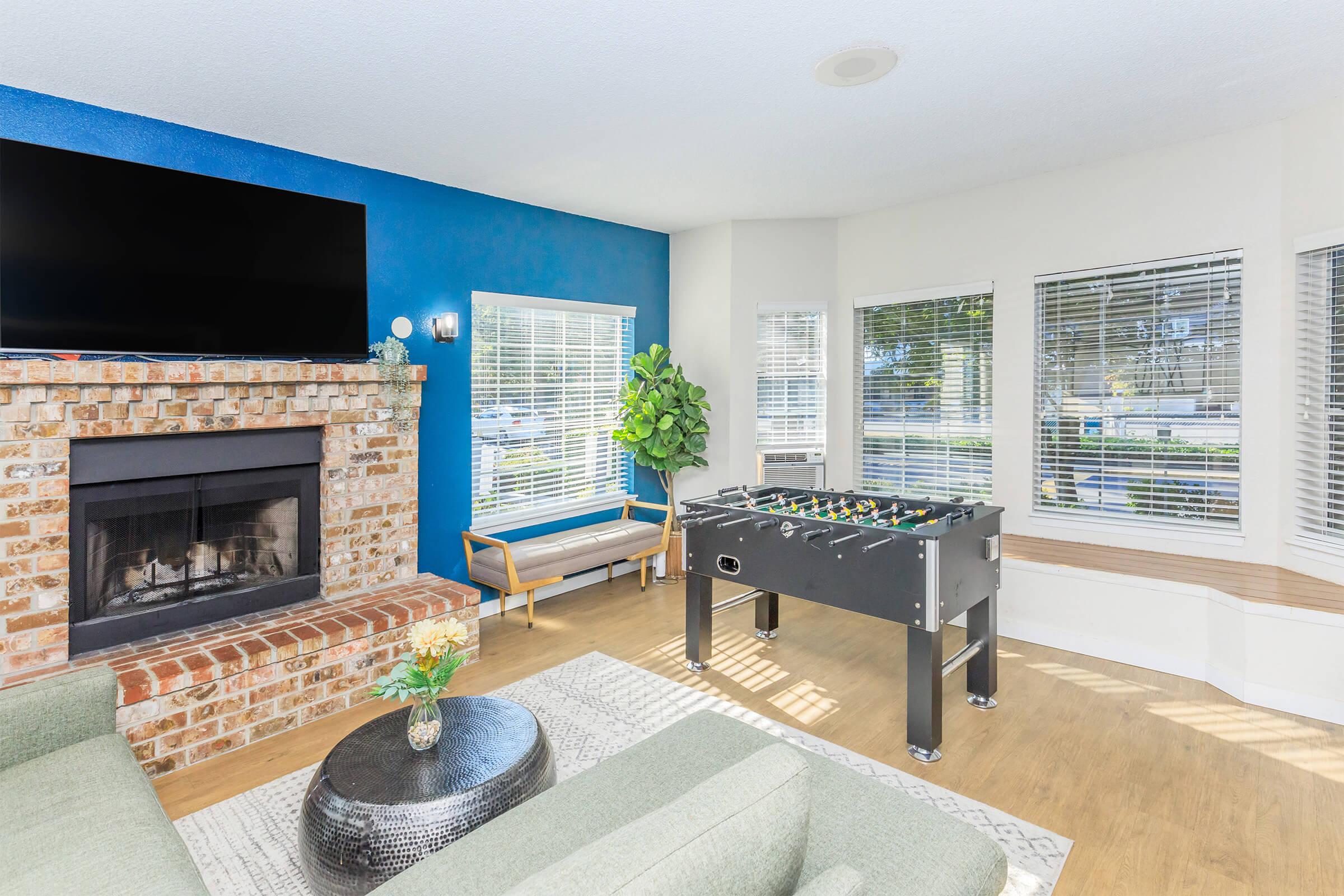 A bright living space featuring a blue accent wall and large windows with white blinds. There is a cozy seating area with a green sofa, a round black coffee table, and a foosball table. A brick fireplace adds warmth, complemented by a potted plant near the windows. Overall, the room has a modern and inviting atmosphere.