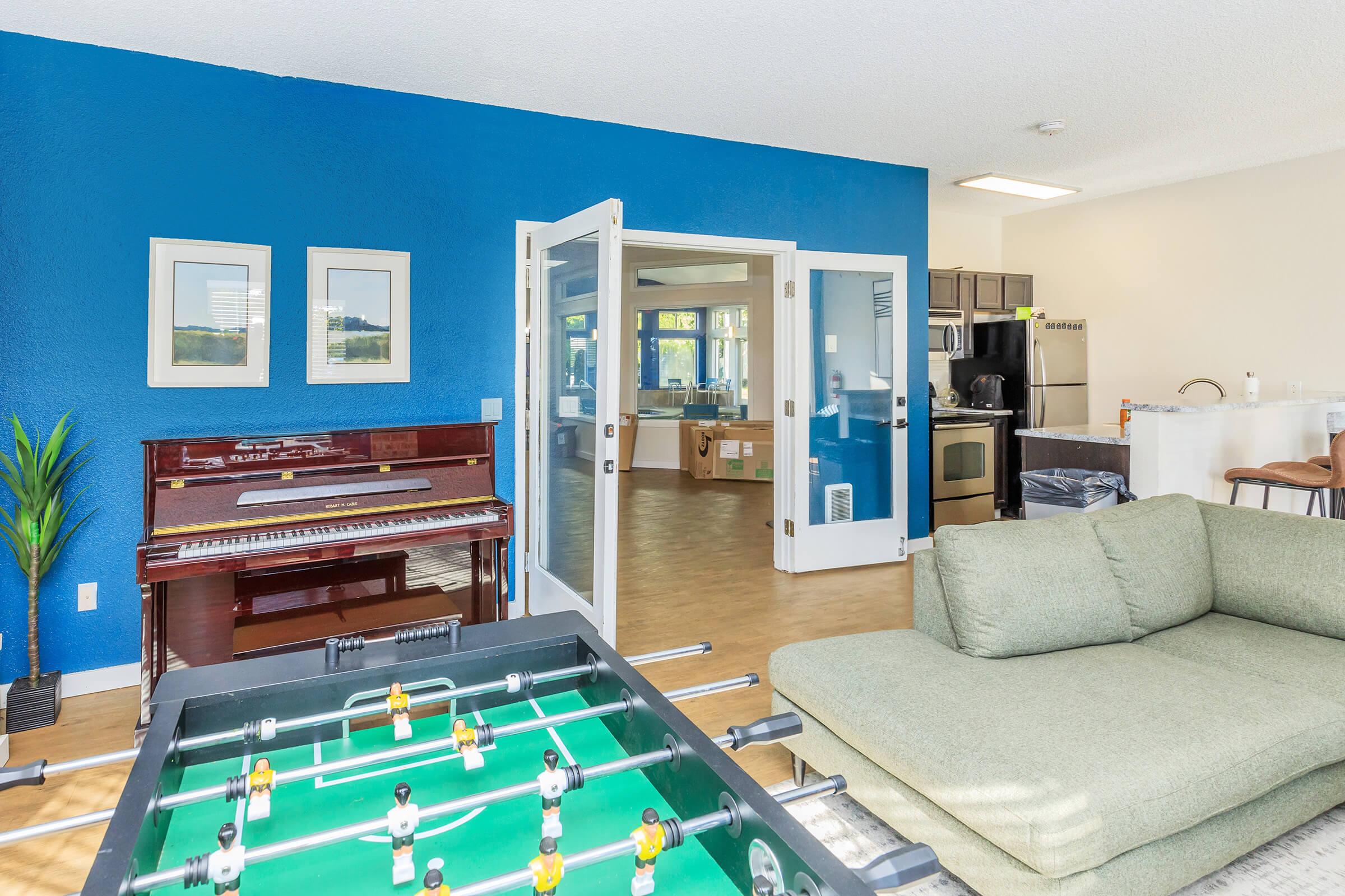 A spacious living area featuring a green sofa, a foosball table, and a vintage piano. The walls are painted blue, and large glass doors lead to a kitchen with modern appliances. Natural light floods the room, creating a bright and inviting atmosphere. A plant adds a touch of greenery to the space.