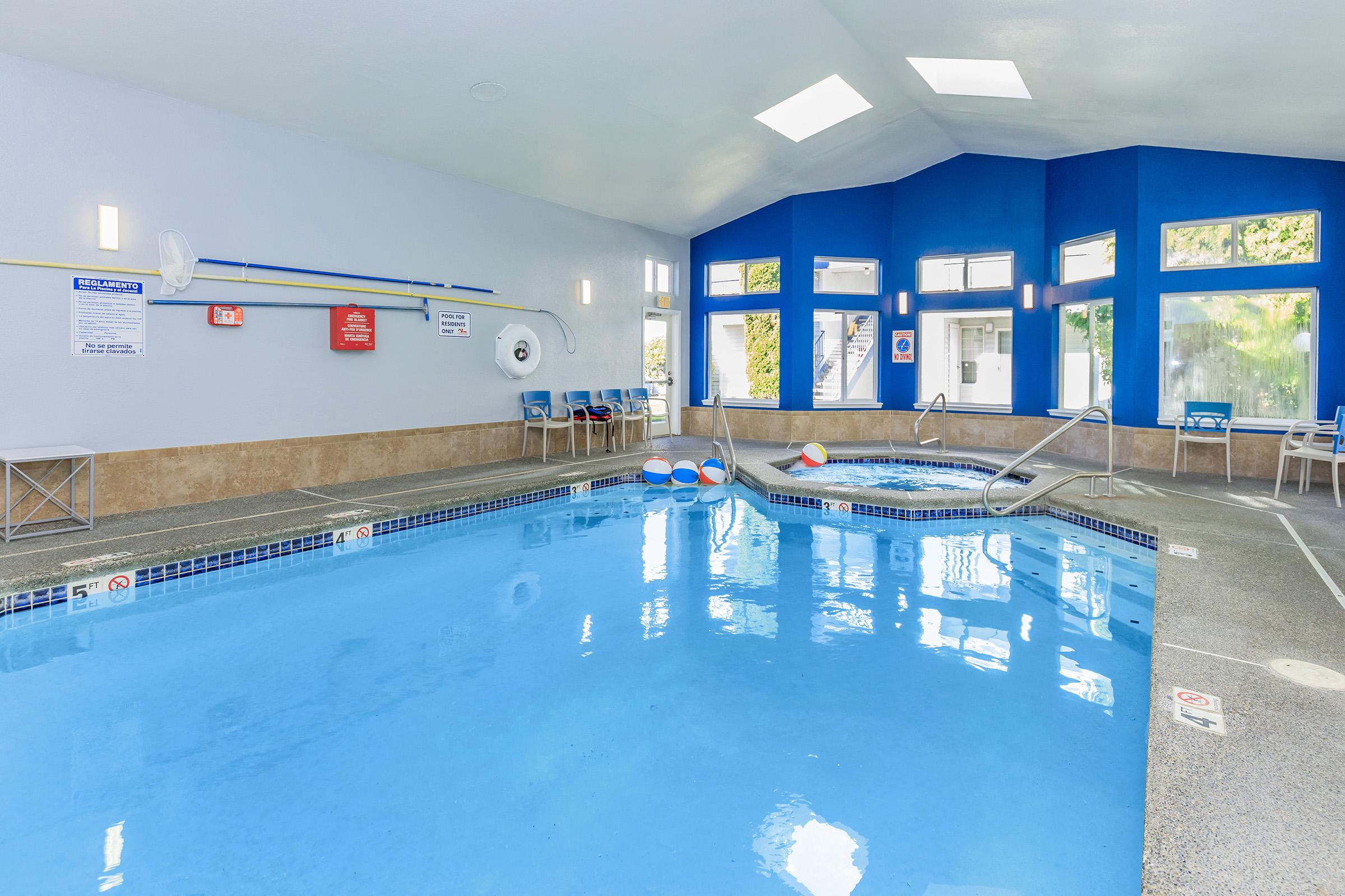 Indoor swimming pool area featuring crystal-clear blue water, a jacuzzi, and bright walls. Large windows allow natural light, and safety equipment is visible on the wall. The space is designed for relaxation and leisure, with chairs positioned around the poolside.