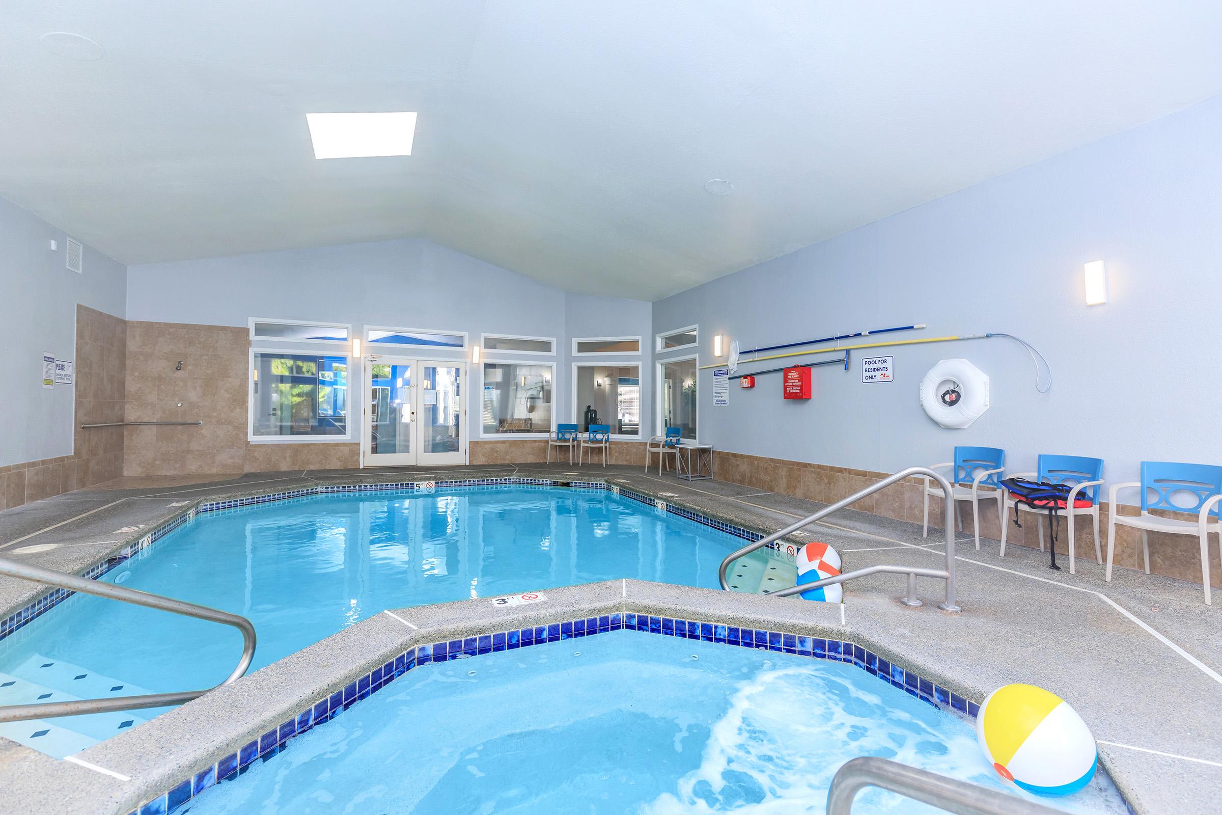 Indoor swimming pool area featuring a hot tub and a shallow pool. The space is bright with large windows, light blue walls, and comfortable seating. Colorful beach balls and safety equipment are visible, creating a welcoming atmosphere for relaxation and fun.