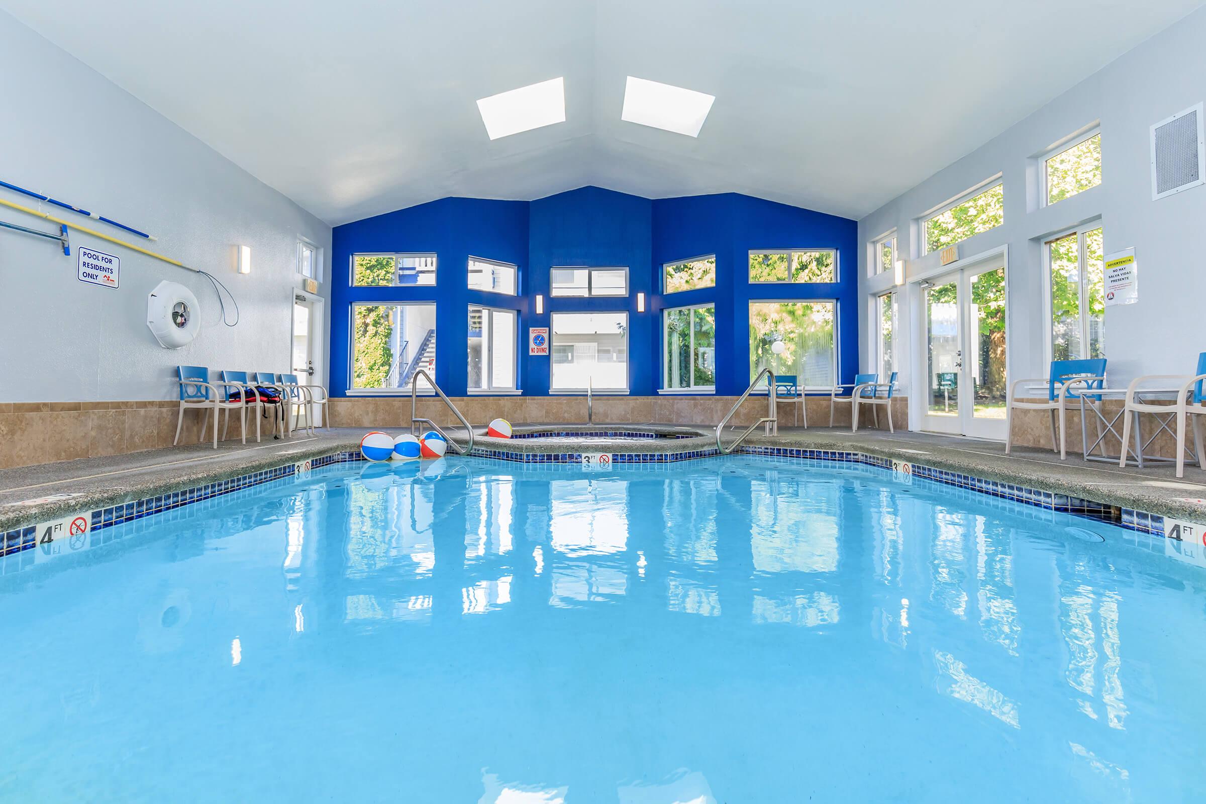 Indoor swimming pool with clear blue water, surrounded by large windows that let in natural light. Colorful pool floats are visible in the water. The walls are painted in shades of blue, and there are lounge chairs arranged around the pool area, creating a relaxing environment.