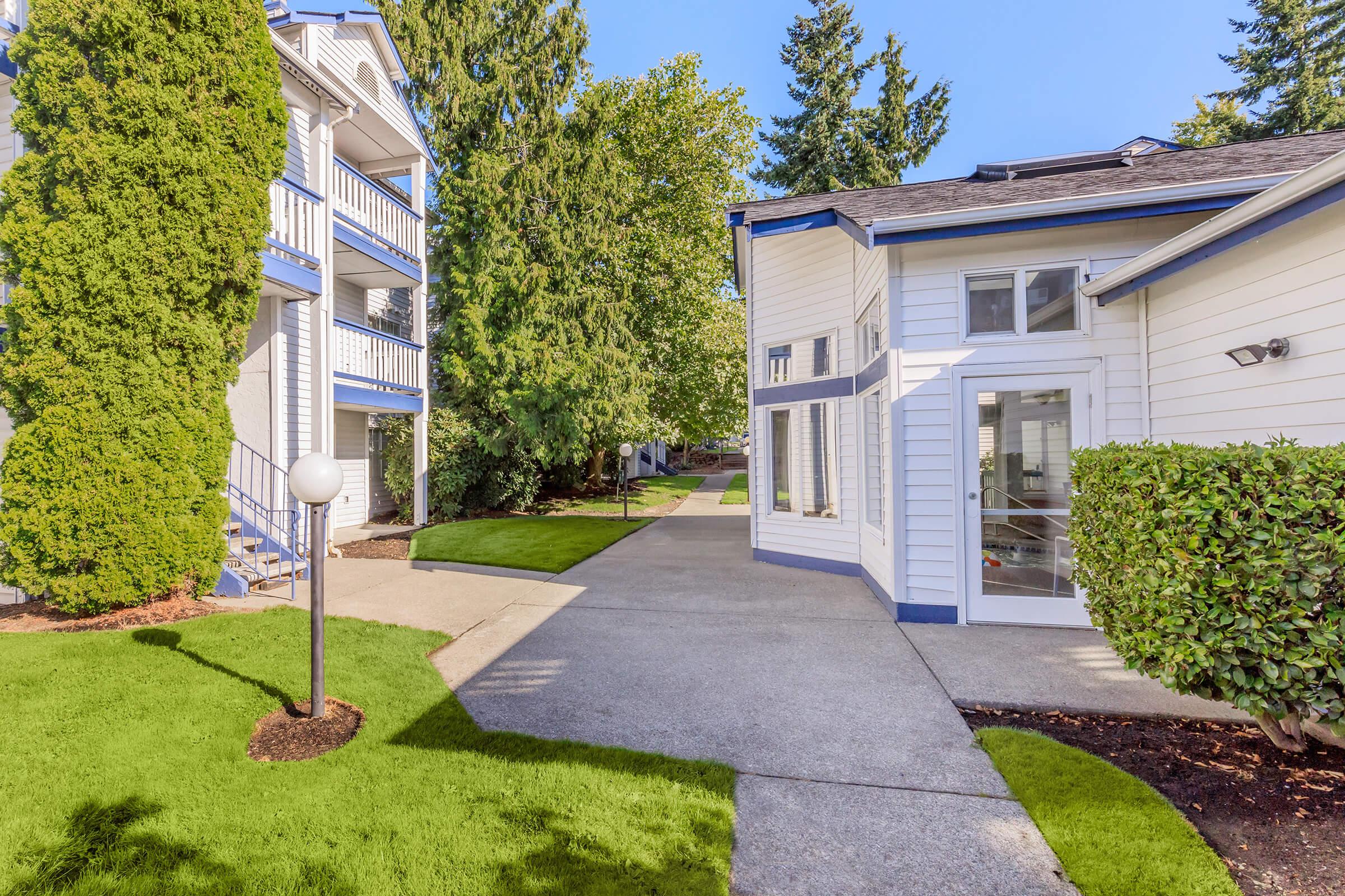 A well-maintained residential complex featuring two buildings with white and blue exteriors, surrounded by green lawns and trees. A pathway leads between the buildings, with a decorative light fixture and shrubbery lining the walkway. Bright sunlight enhances the inviting atmosphere.