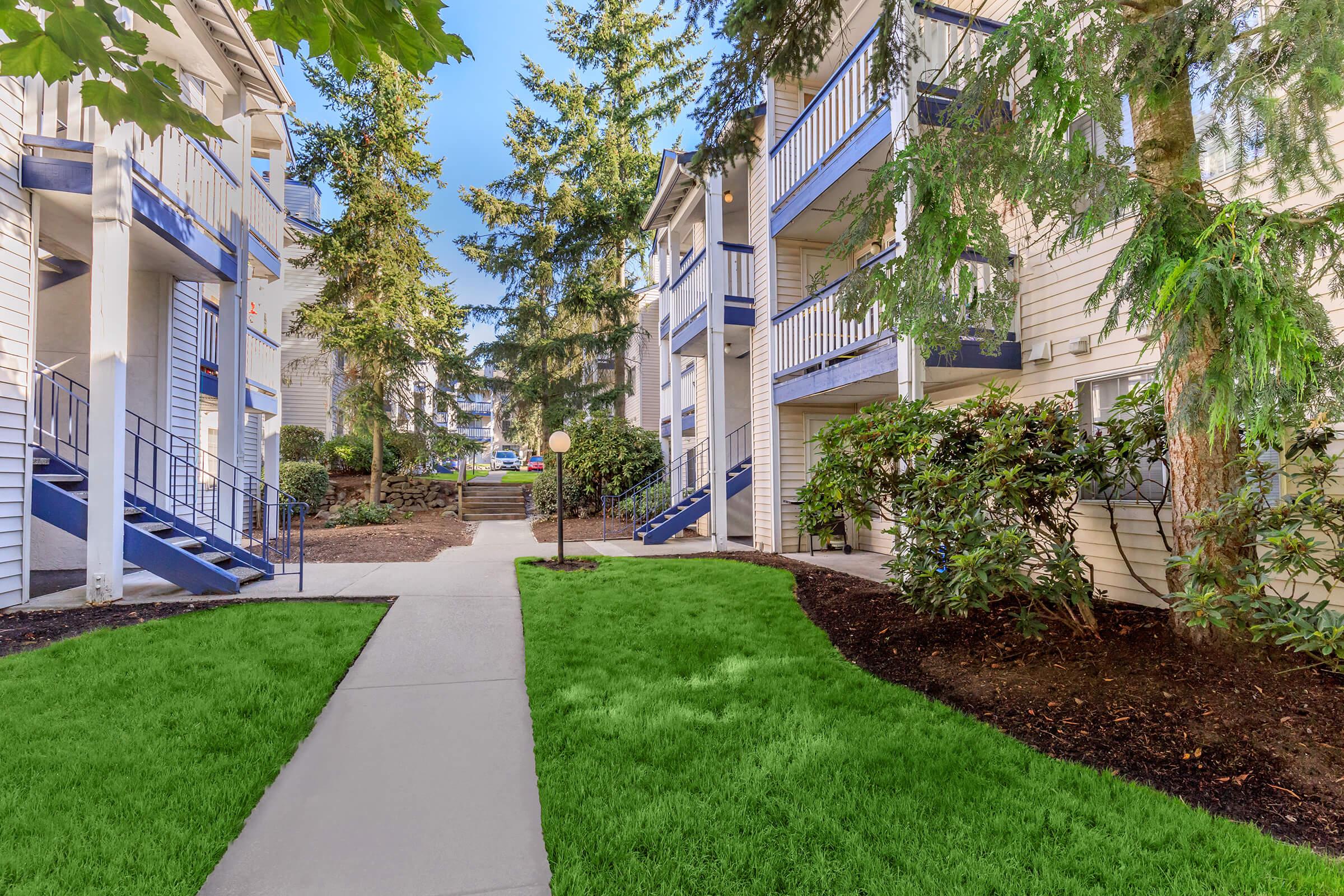 A well-maintained apartment complex featuring a pathway lined with lush green grass, surrounded by trees and landscaped shrubs. The buildings are two to three stories high, with blue railings and staircases leading to individual units. Soft natural lighting enhances the welcoming atmosphere of the outdoor area.