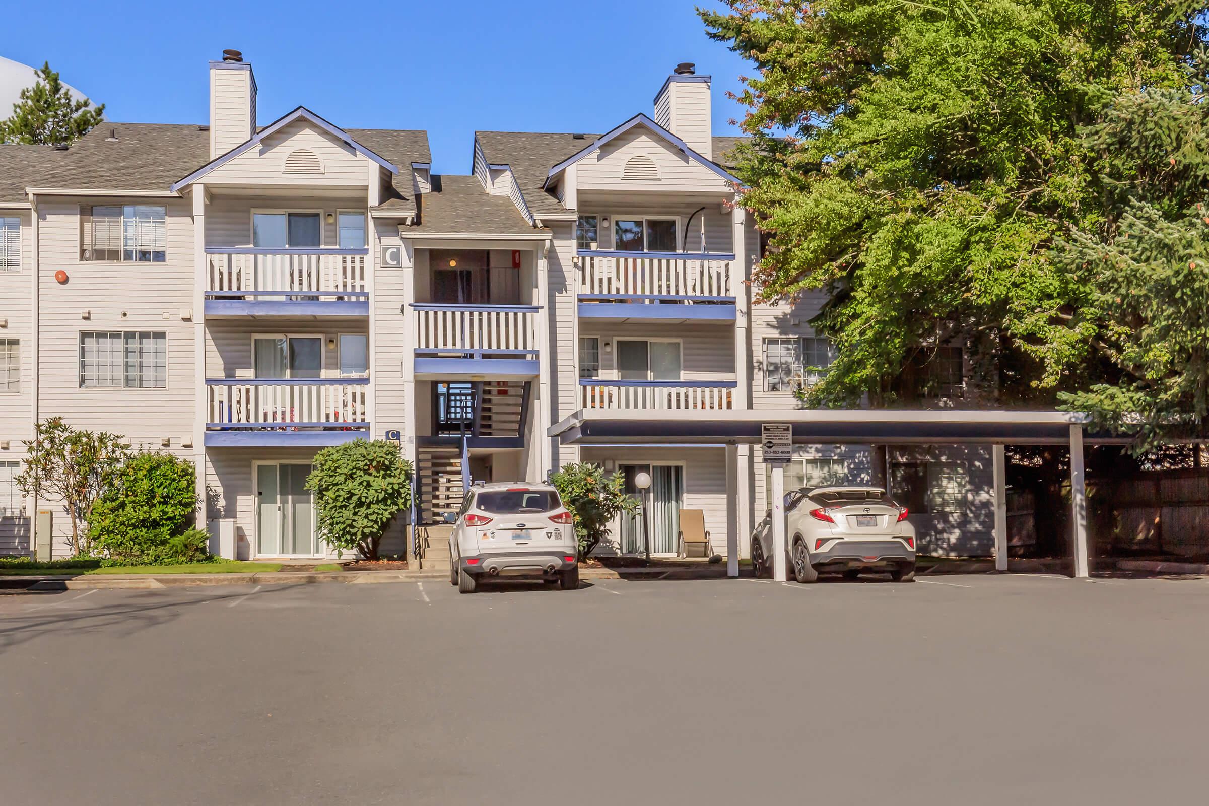 A residential building with three stories featuring balconies, surrounded by greenery. Two parked cars are visible in the foreground, and a covered parking area is located to the right. The sky is clear and blue, indicating pleasant weather.
