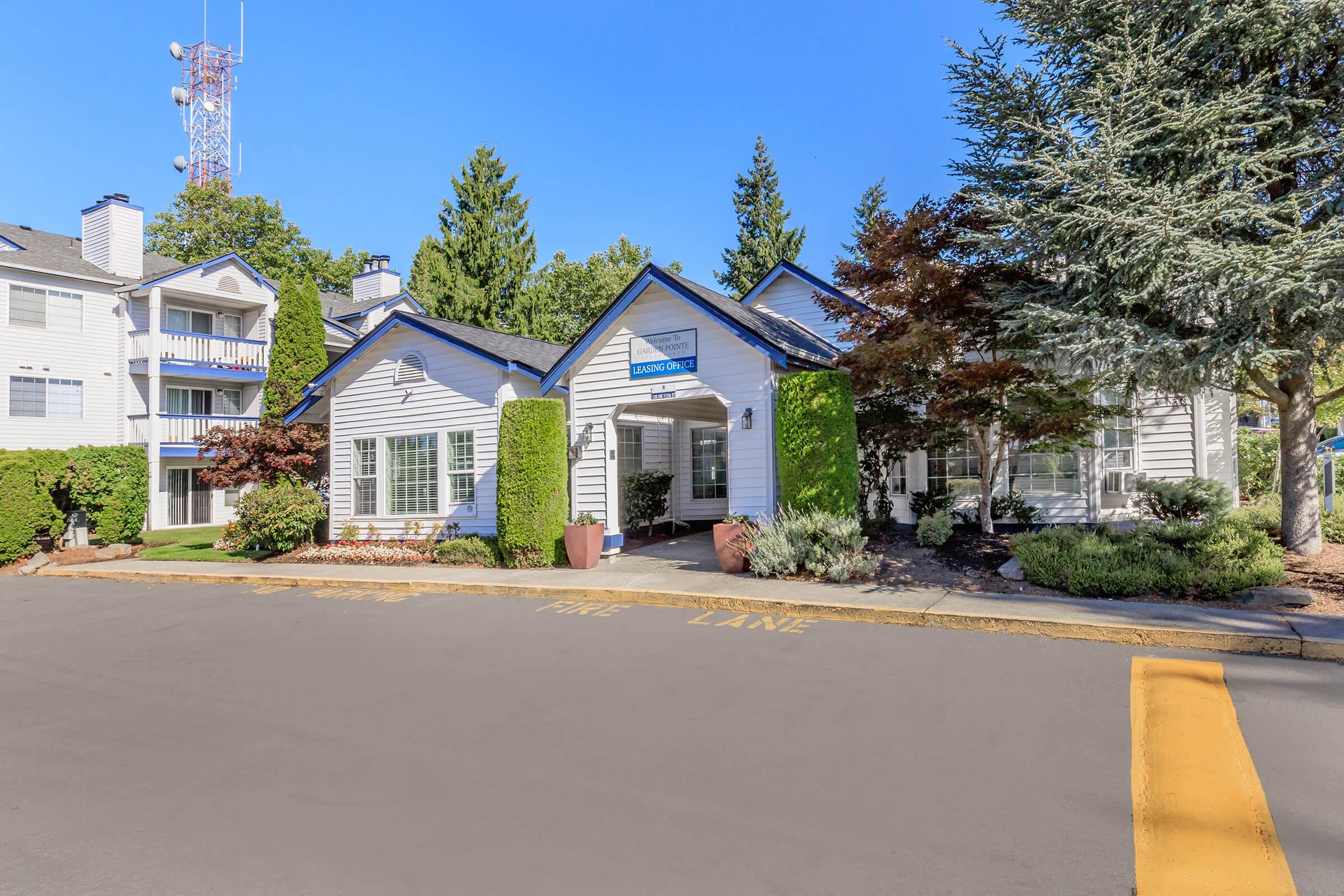 A well-maintained entrance to a residential building featuring a white exterior, surrounded by trees and shrubs. The driveway in front has a yellow-painted line, and a sign indicates the building’s name or purpose. Clear blue sky overhead enhances the inviting atmosphere of the location.