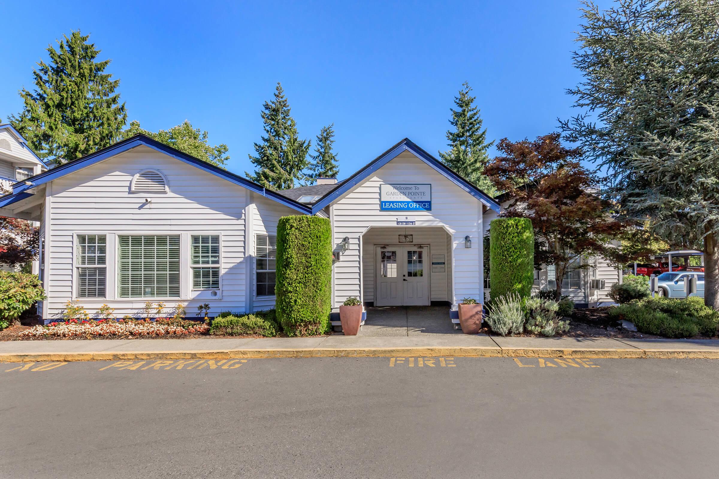 A single-story building with a welcoming entrance, featuring a sign that says "Leasing Office." Surrounding the building are well-maintained plants and trees, with a clear blue sky overhead. The pathway is marked for parking and fire lane, adding to the organized appearance of the property.