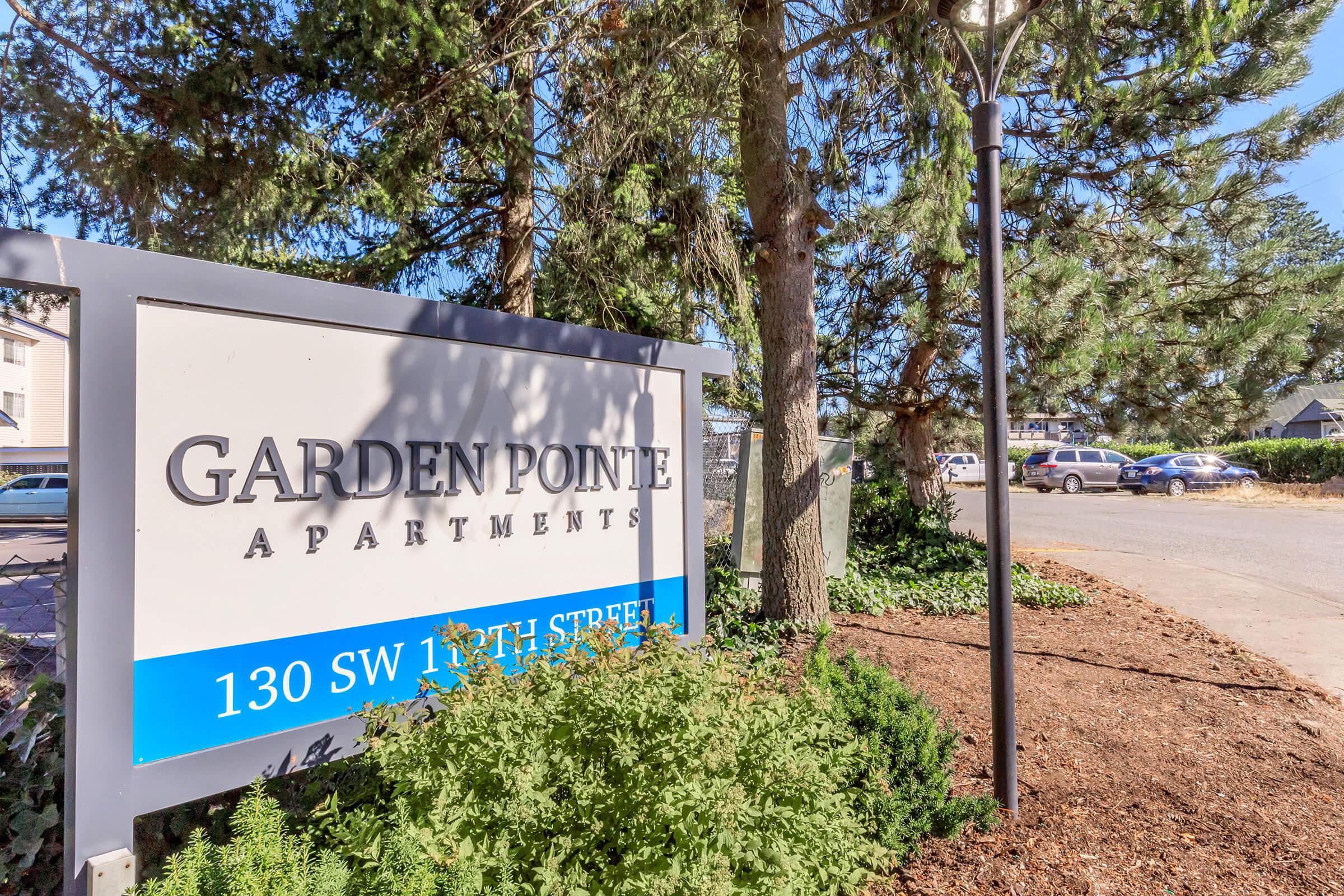Sign for Garden Pointe Apartments located at 130 SW 13th Street. The sign features a modern design and is surrounded by greenery, including trees and shrubs. Parking spaces are visible in the background.