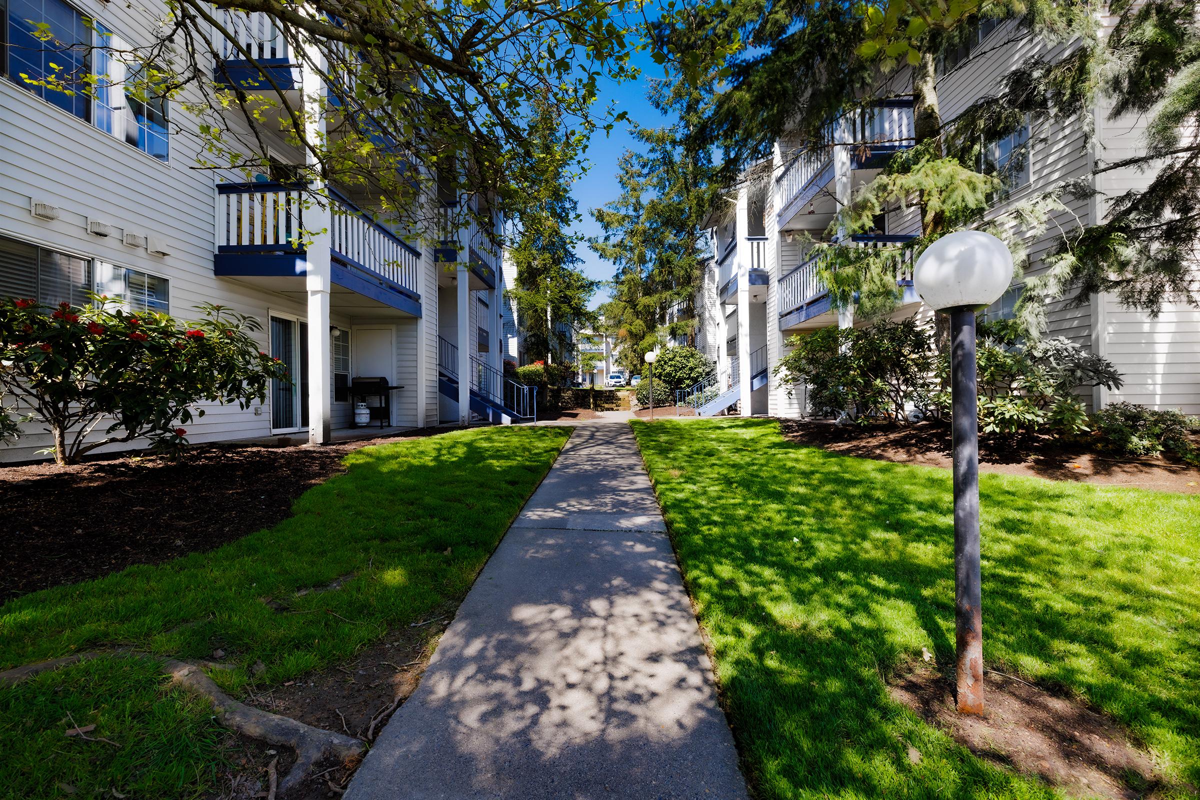 A well-maintained apartment complex featuring two-story buildings lined with balconies. A paved pathway runs through a lush green lawn, surrounded by decorative shrubs and trees. The scene is bright and sunny, with clear blue skies overhead.