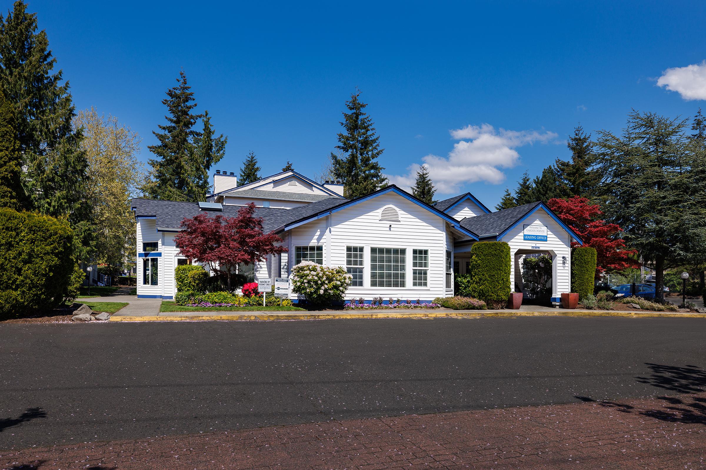 A charming white house with blue accents, surrounded by lush greenery and colorful flowering plants. Tall trees frame the scene under a bright blue sky with a few clouds. The driveway is paved, leading to the house, which features large windows and a welcoming entrance.
