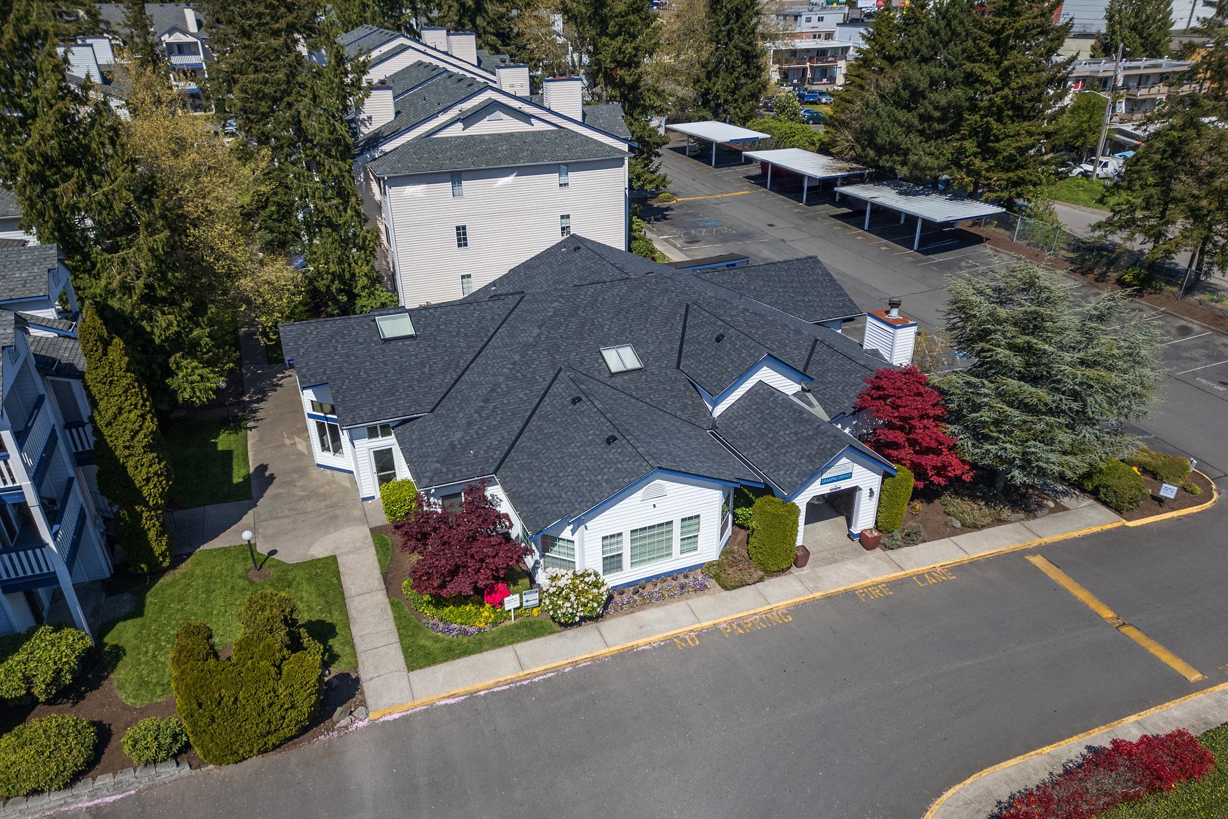 Aerial view of a multi-unit residential building surrounded by trees and landscaping. The building features a dark roof and light-blue siding, with prominent red and green bushes in the foreground. There is a parking area visible in the background with designated no parking signs.