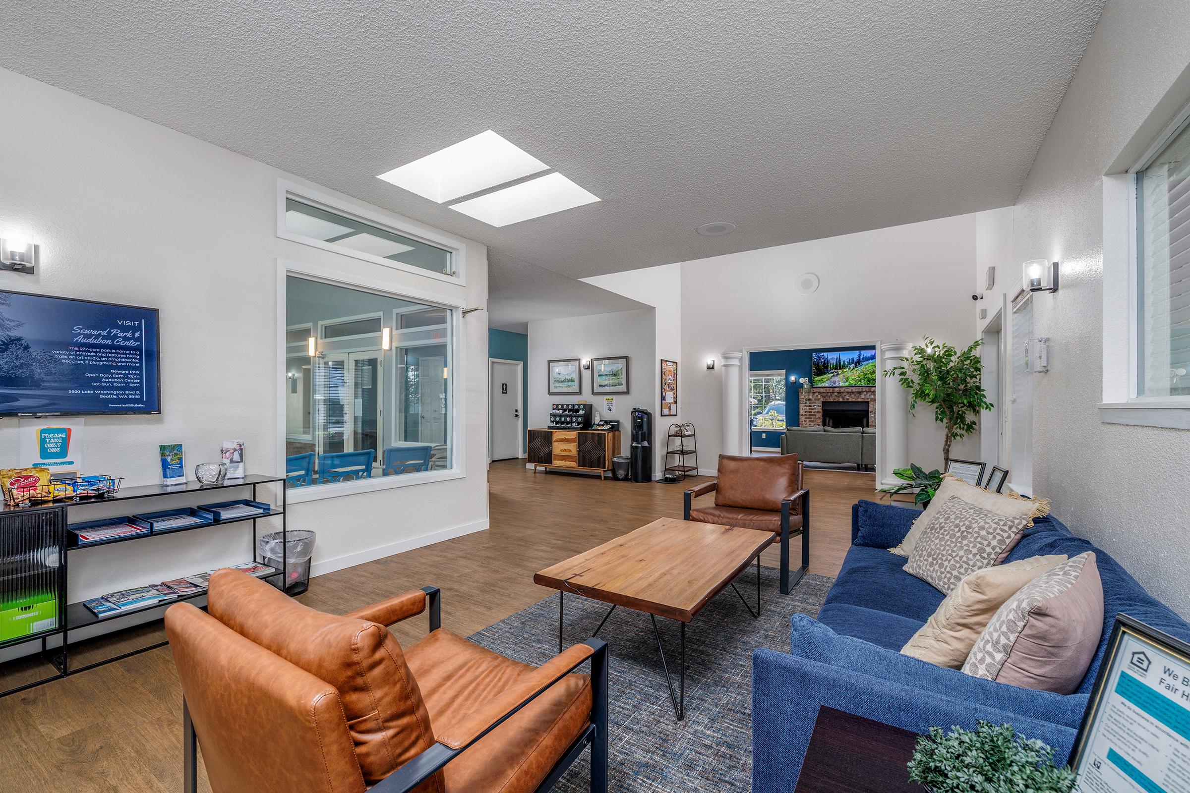 A modern, bright common area featuring comfortable seating with brown and blue chairs, a wooden coffee table, and a large window. A kitchenette is visible in the background, along with decorative plants and wall art, creating a welcoming atmosphere for residents or guests. Bright lighting enhances the open space.