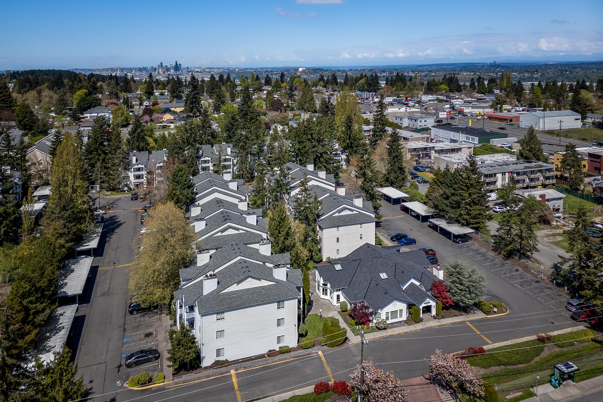 Aerial view of a suburban area featuring residential buildings surrounded by trees, with a city skyline visible in the background. The scene includes parking lots and commercial properties, showcasing a blend of nature and urban infrastructure on a clear day.