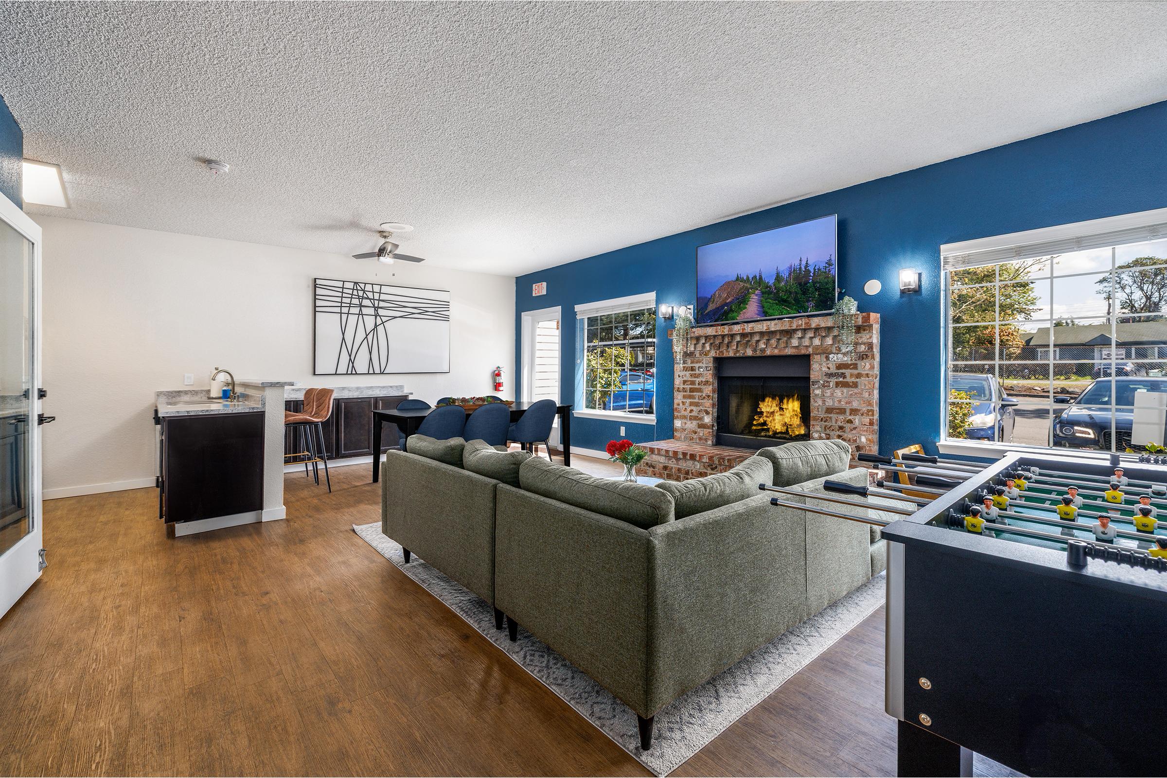A cozy living room featuring a green sofa, a fireplace with a television above, and a modern wall art piece. There's a dining area with chairs and a kitchen counter on one side, along with a foosball table. Sunlight streams through large windows, creating a warm and inviting atmosphere.