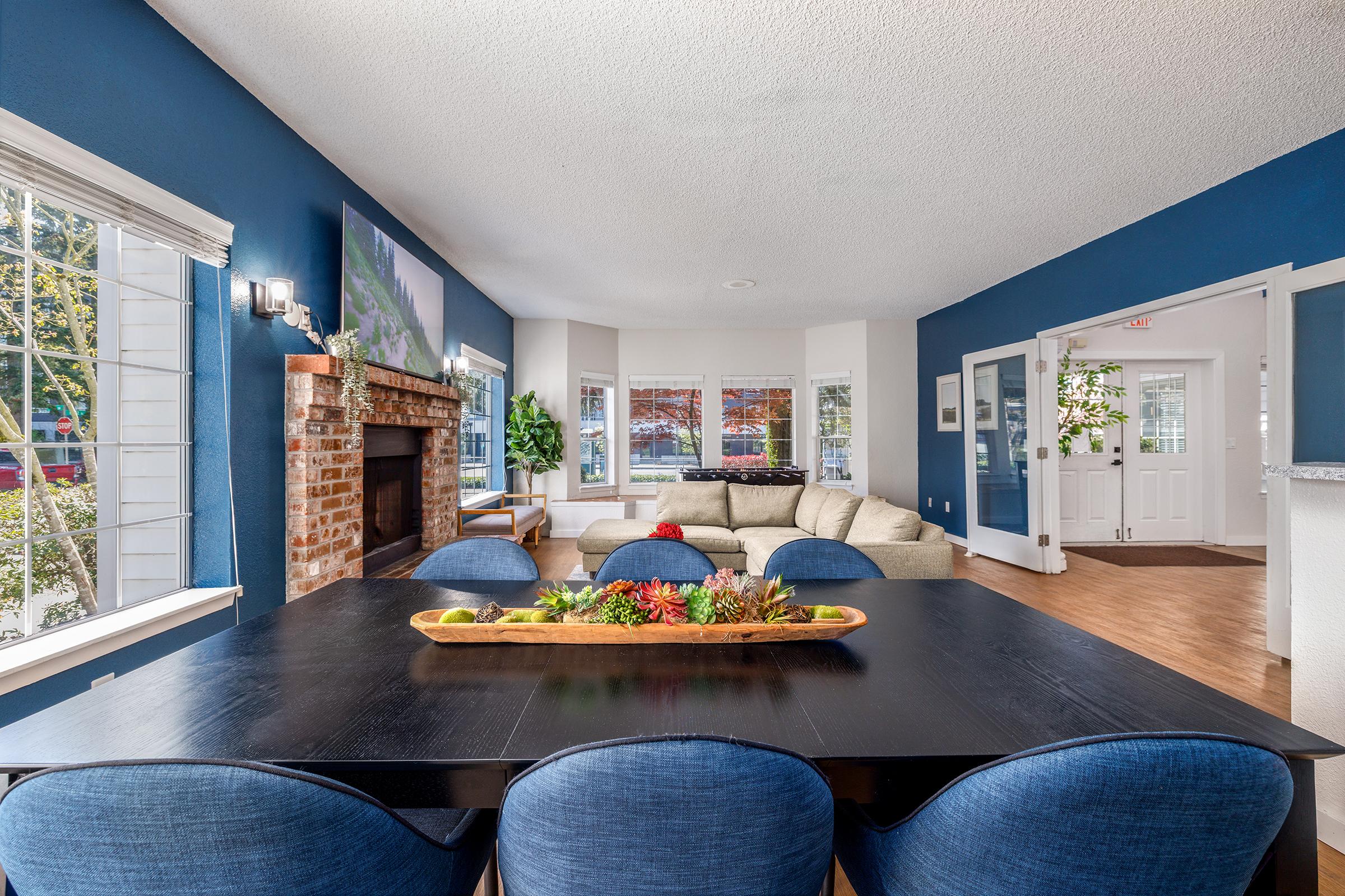 A cozy living room with a dark blue accent wall, featuring a dining table with blue upholstered chairs. A wooden centerpiece holds decorative plants. There is a fireplace with a brick surround, large windows letting in natural light, and a plush sofa in the corner, all creating a welcoming atmosphere.