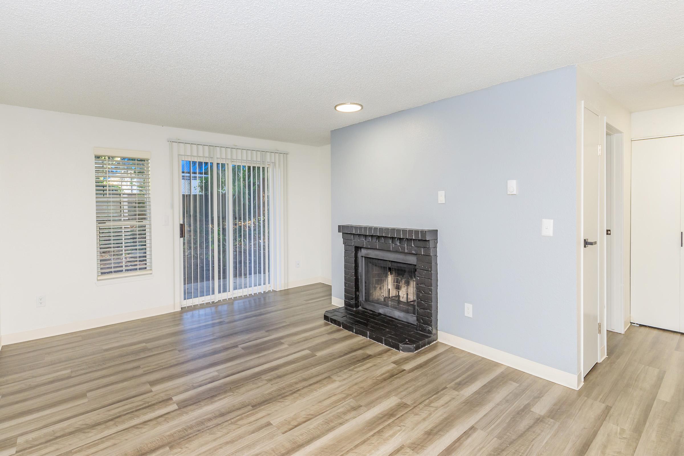 A spacious, light-filled room featuring a modern fireplace, large windows with vertical blinds, and light wood-style flooring. The walls are painted in soft blue and white, enhancing the inviting atmosphere. A doorway leads to another room, with a sliding glass door opening to an outdoor area.