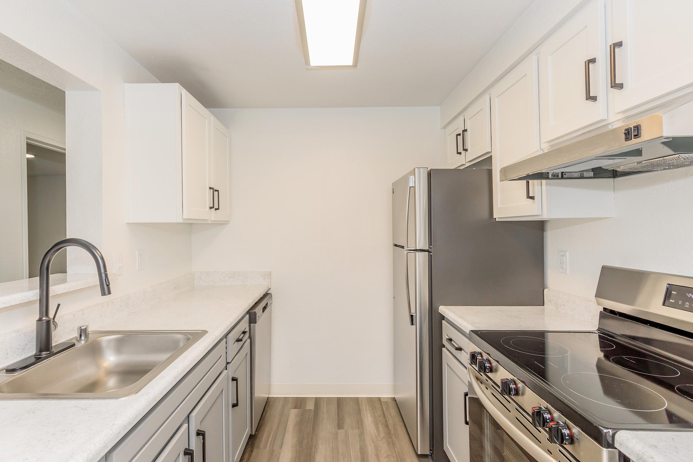 Modern kitchen with white cabinets, stainless steel appliances including a refrigerator and an electric stove, a stainless steel sink, and light-colored countertops. The space is well-lit with a ceiling fixture and features a sleek, contemporary design with a neutral color palette.