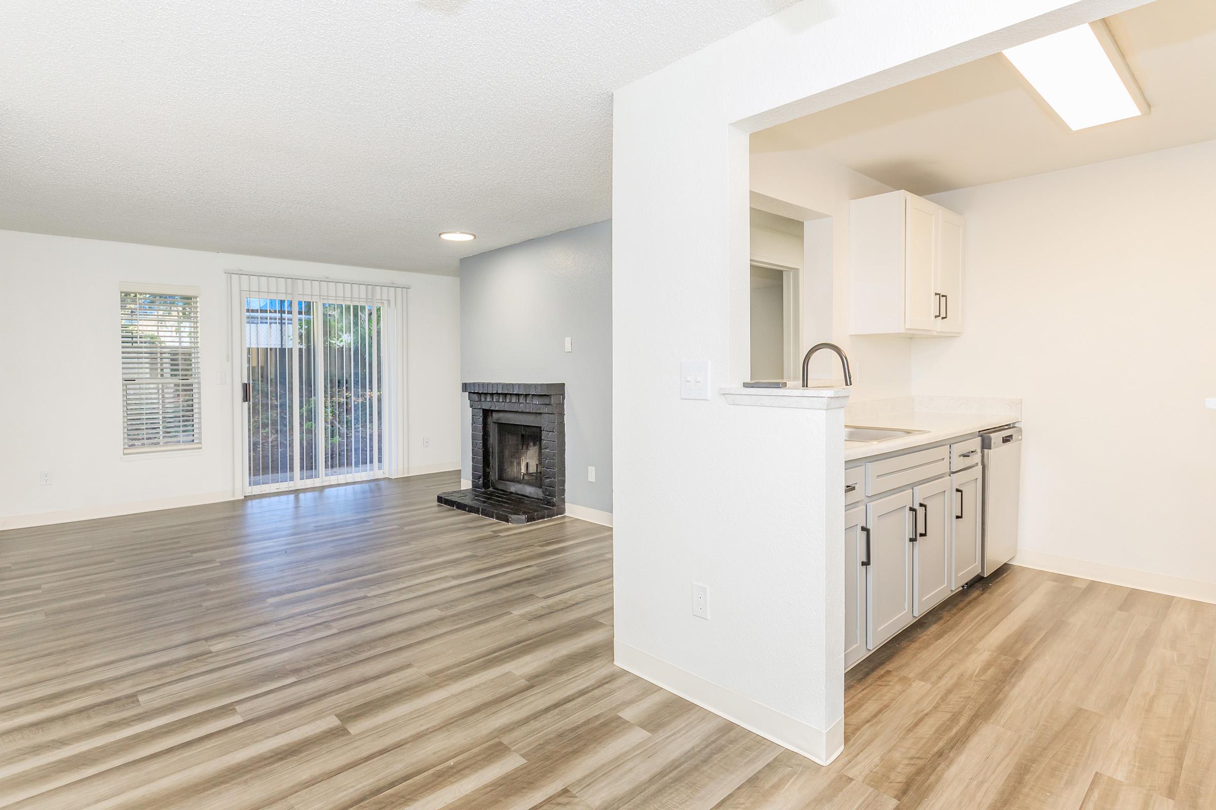 Spacious living area featuring a modern kitchen with stainless steel appliances, a black fireplace, and large windows with natural light. The room is designed with light wood flooring, white walls, and a sliding glass door leading to an outdoor space.
