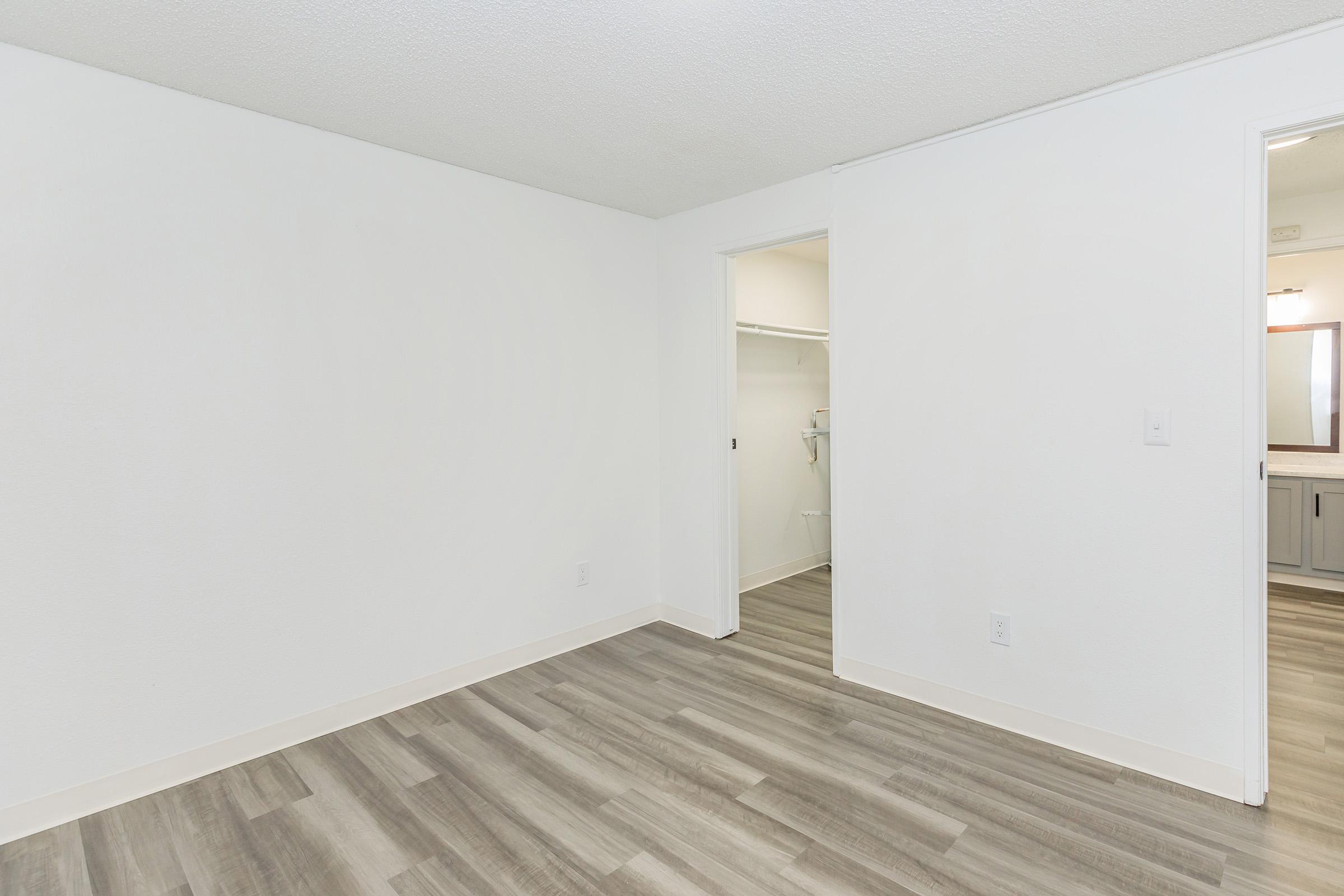 Empty room with white walls and light wood laminate flooring. A doorway leads to a small closet on the left, while a second doorway on the right opens into a bathroom area. Natural light may be coming from an unseen window, creating a bright and spacious atmosphere.