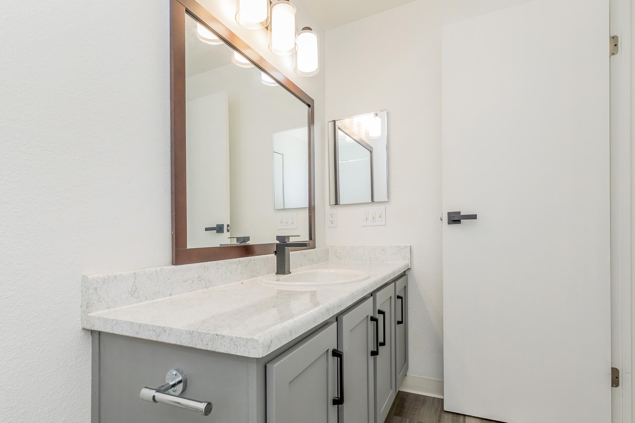 A modern bathroom featuring a gray vanity with a white countertop and a sink. Above the vanity, there are two mirrors and stylish light fixtures. A clean, minimalist aesthetic with light walls and a white door visible on the right. The floor is a light wood laminate.
