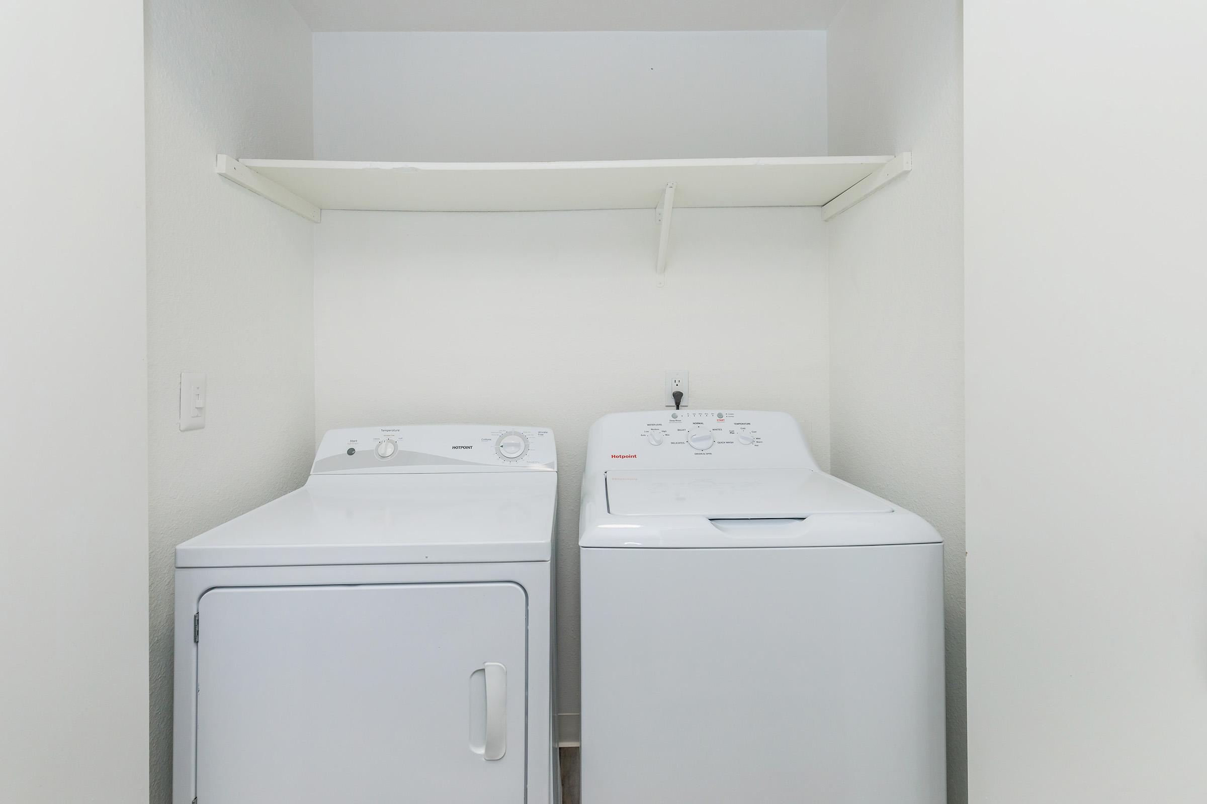 A laundry room featuring a white washer and dryer side by side against a plain white wall. Above them is a simple shelf for storage. The space is well-lit and minimalistic, emphasizing the appliances and functional design.