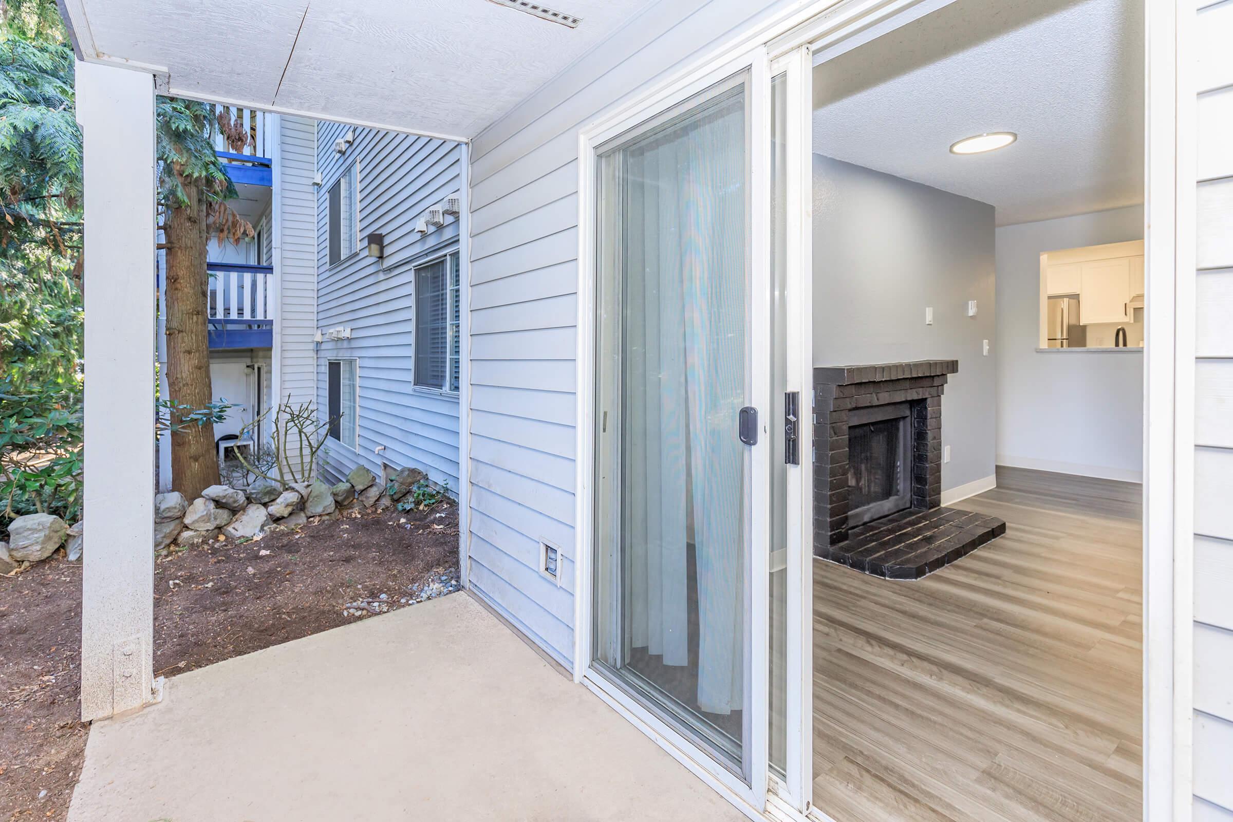 A view of a patio entrance with sliding glass doors leading into a room featuring a black fireplace. The exterior has light blue siding and greenery in the background, while the interior appears modern with wood-like flooring and neutral wall colors.