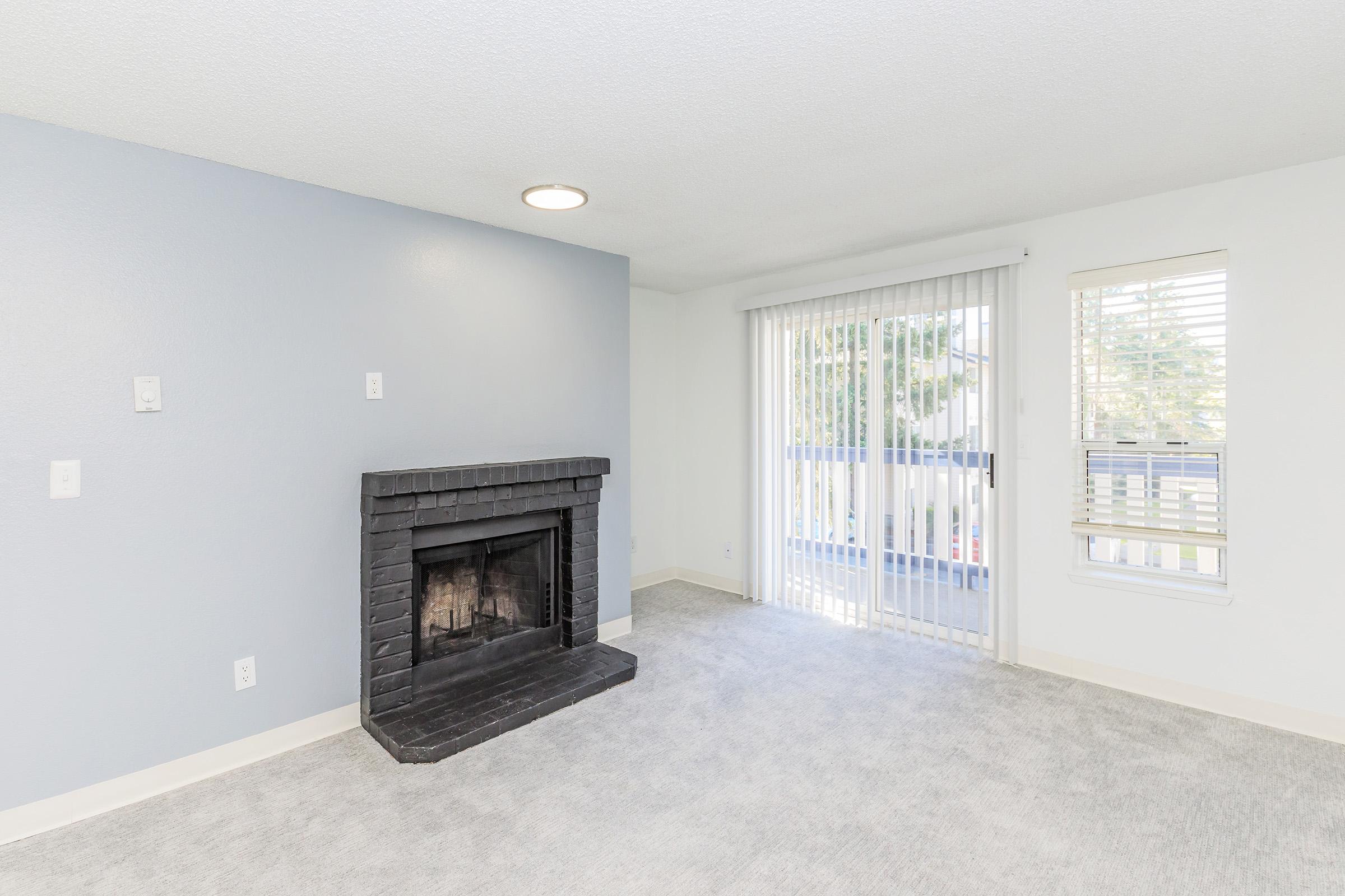 A cozy interior space featuring a light gray carpet, a black brick fireplace, and a wall painted in a soft blue hue. Large windows with vertical blinds allow natural light to fill the room. The area is uncluttered and ready for furniture placement, creating an inviting living atmosphere.