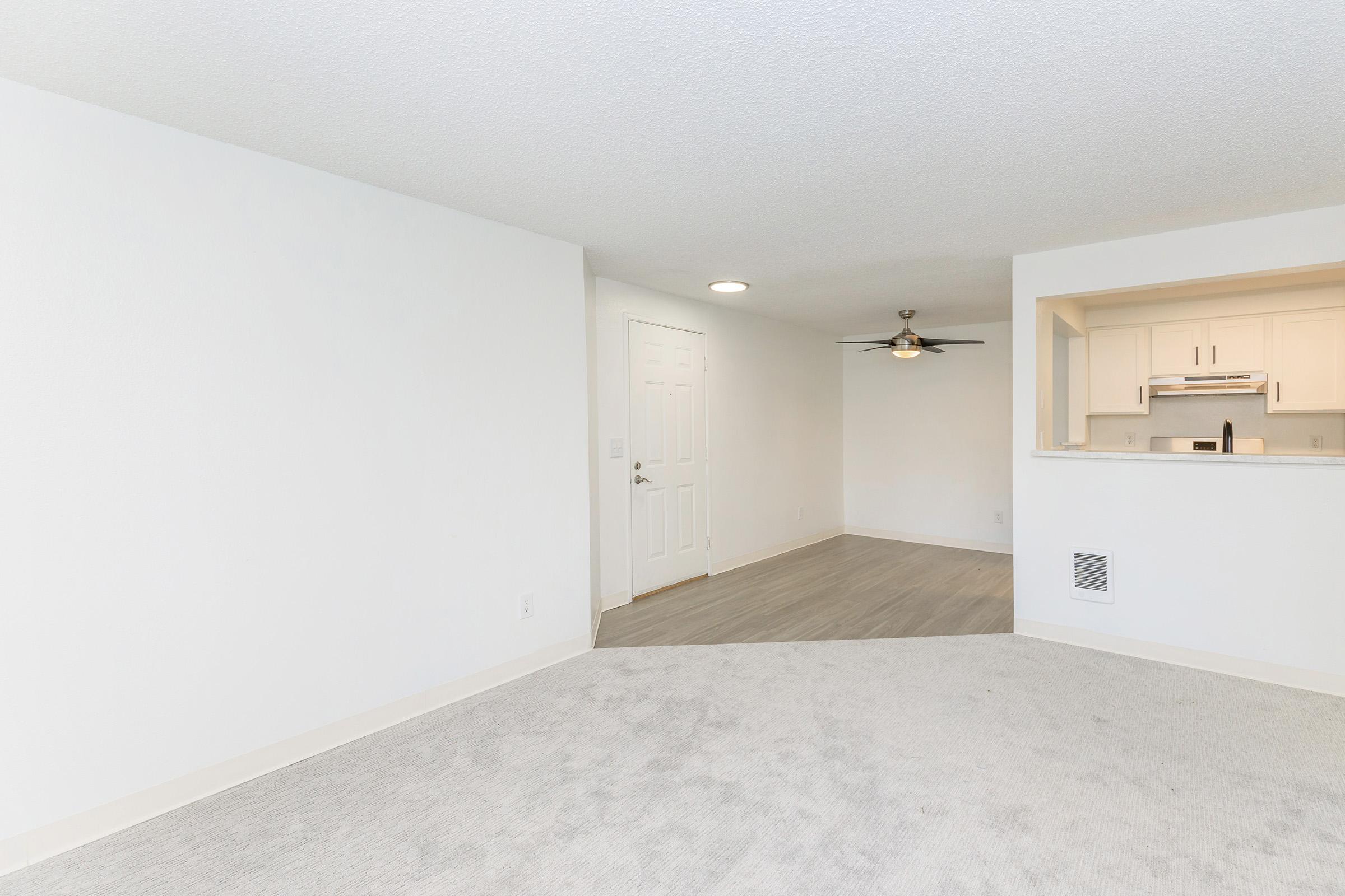 An empty, well-lit room with light-colored walls and carpet. A doorway is visible on one side, leading to another room. A ceiling fan is mounted in the adjacent area, which has a view into a kitchen or dining space. The overall atmosphere is clean and modern, suitable for various uses.