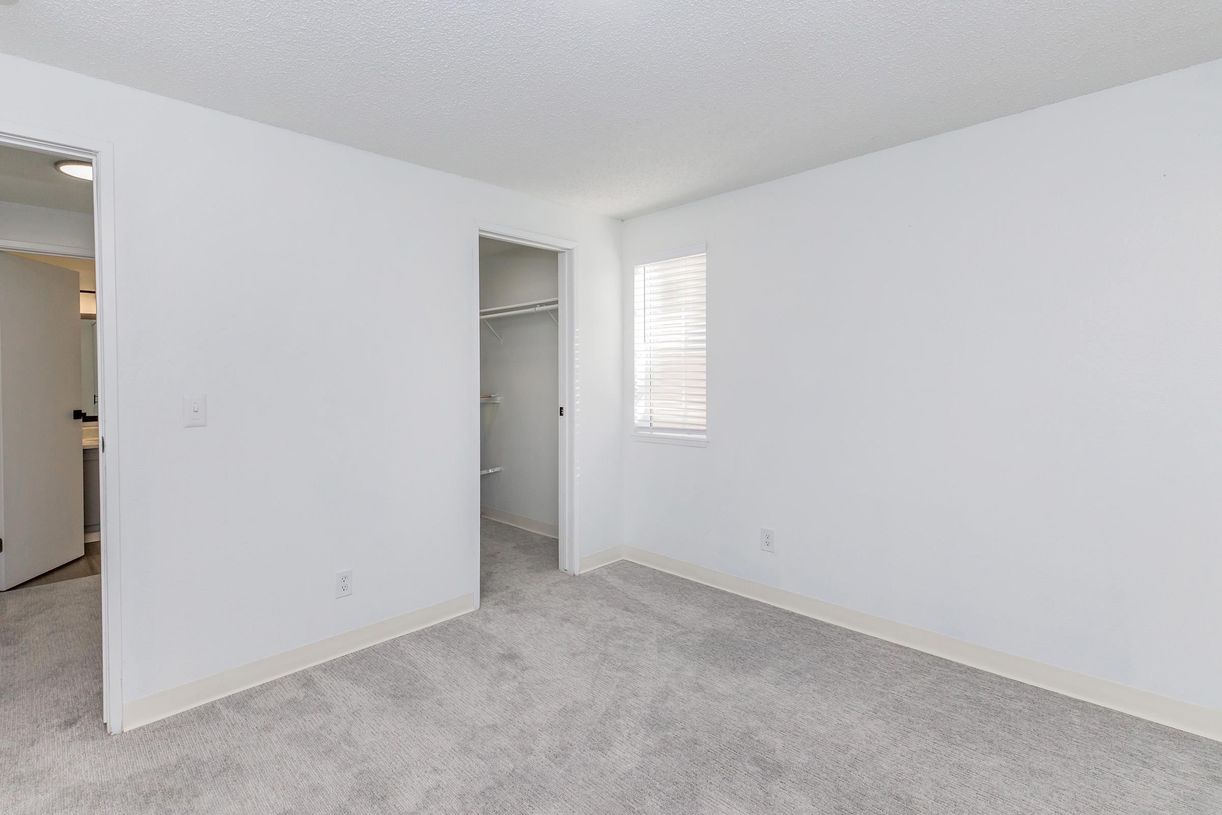 A bright, empty bedroom featuring light gray carpet and white walls. There is a small window with blinds allowing natural light in, and a closet with open doors. The room is unfurnished and spacious, providing a clean and simple aesthetic.