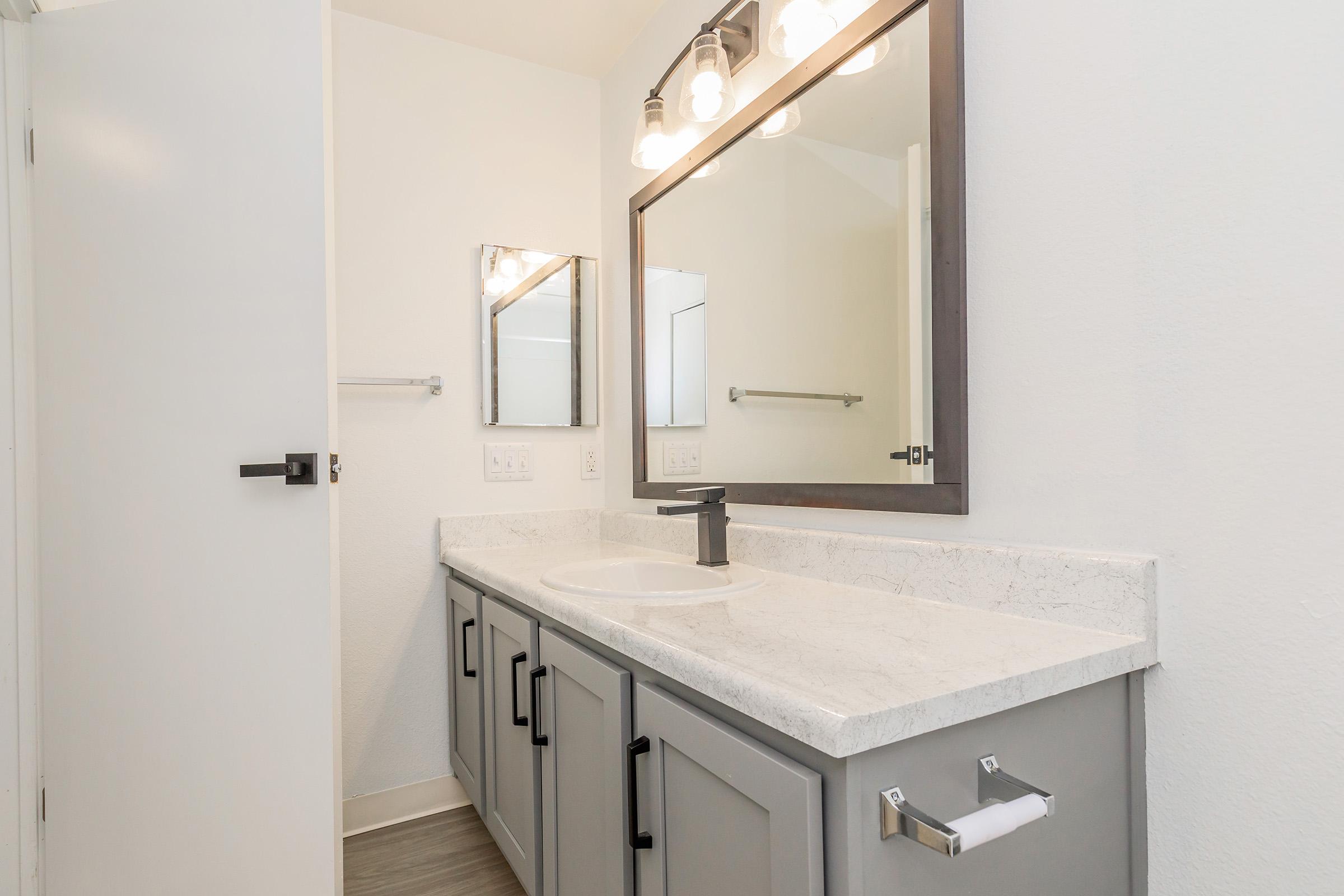 A modern bathroom featuring a gray vanity with a marble countertop, a round sink, and a large mirror with bright lights above. There's a white wall and a door, with a towel bar mounted beside the mirror. Natural light comes in from an unseen window, creating a bright and airy feel.