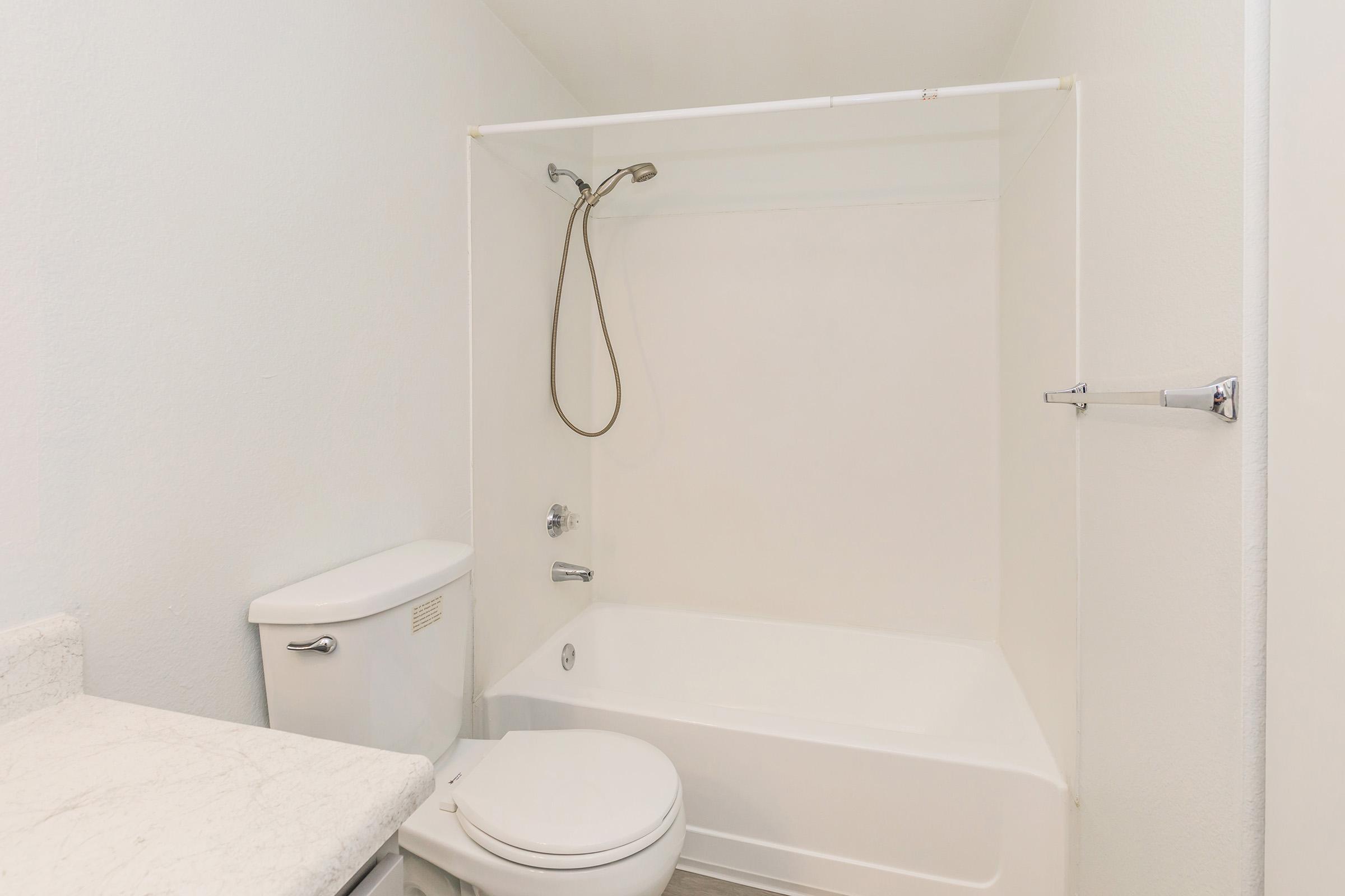 A clean, simple bathroom featuring a bathtub with a showerhead, a white toilet, and a light-colored countertop. The walls are painted a neutral color, creating a bright and spacious feel. The space includes a towel bar on the wall.