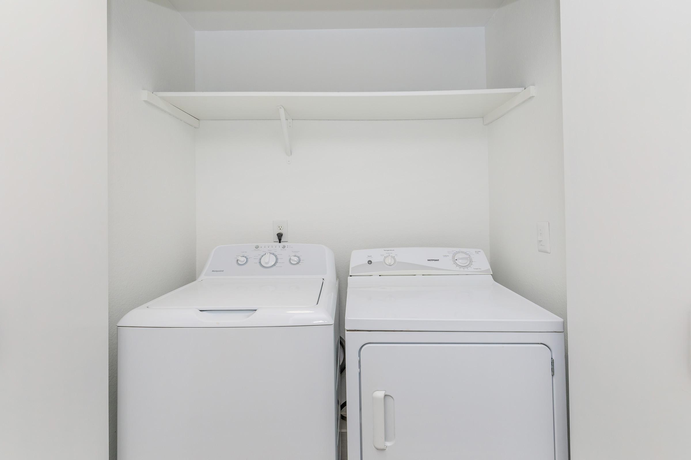 A clean laundry area featuring a washing machine and a dryer, both in white. The appliances are positioned side by side against a plain wall, with a shelf overhead providing additional storage space. The environment is well-lit and organized, suitable for household tasks.