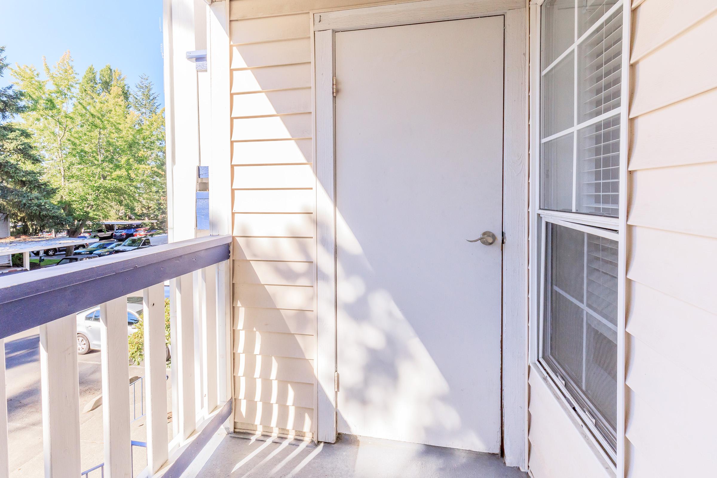A close-up view of a white door on a light-colored apartment balcony. The door is framed by beige siding, with sunlight casting shadows on the wall. A glimpse of trees and parked cars can be seen in the background through a window.