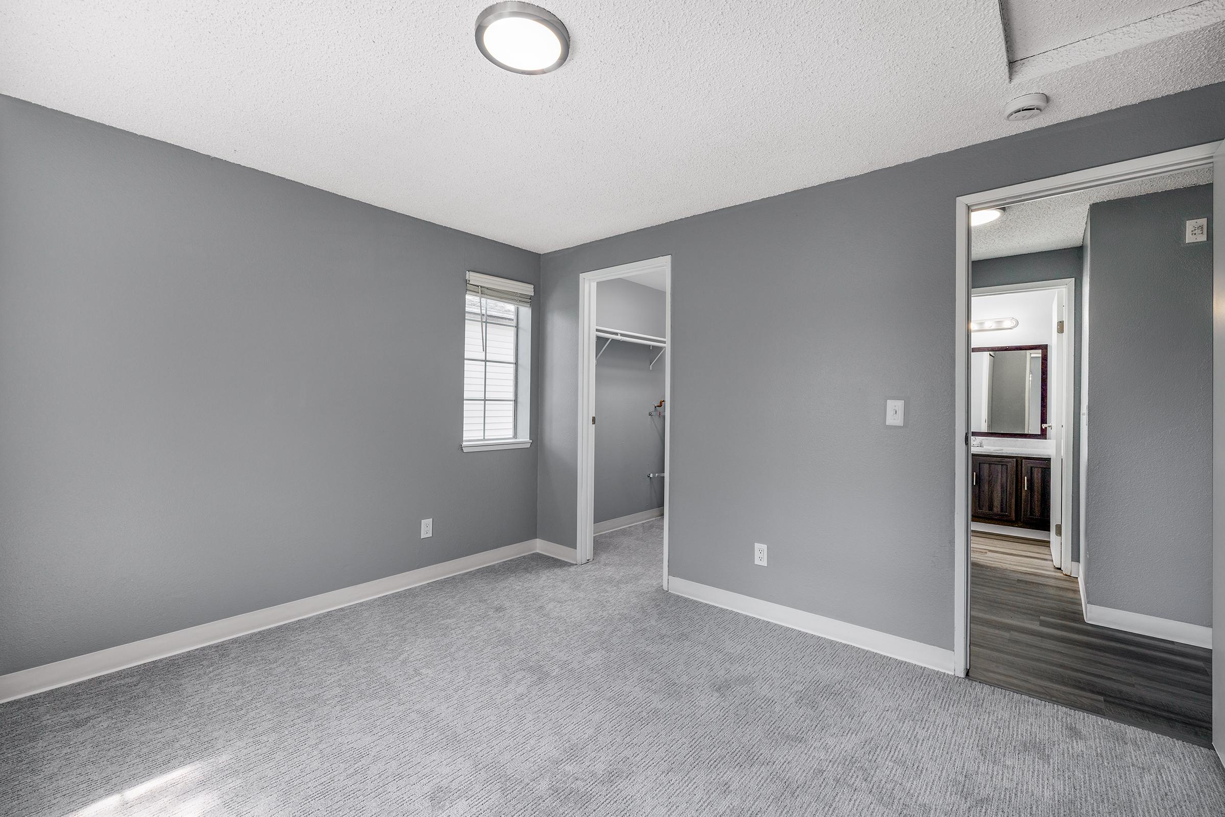 A spacious, empty room with gray walls and carpet. There's a small window on the left, a door leading to a bathroom in the back, and an open closet on the right. The lighting is soft, with a ceiling light fixture illuminating the area.