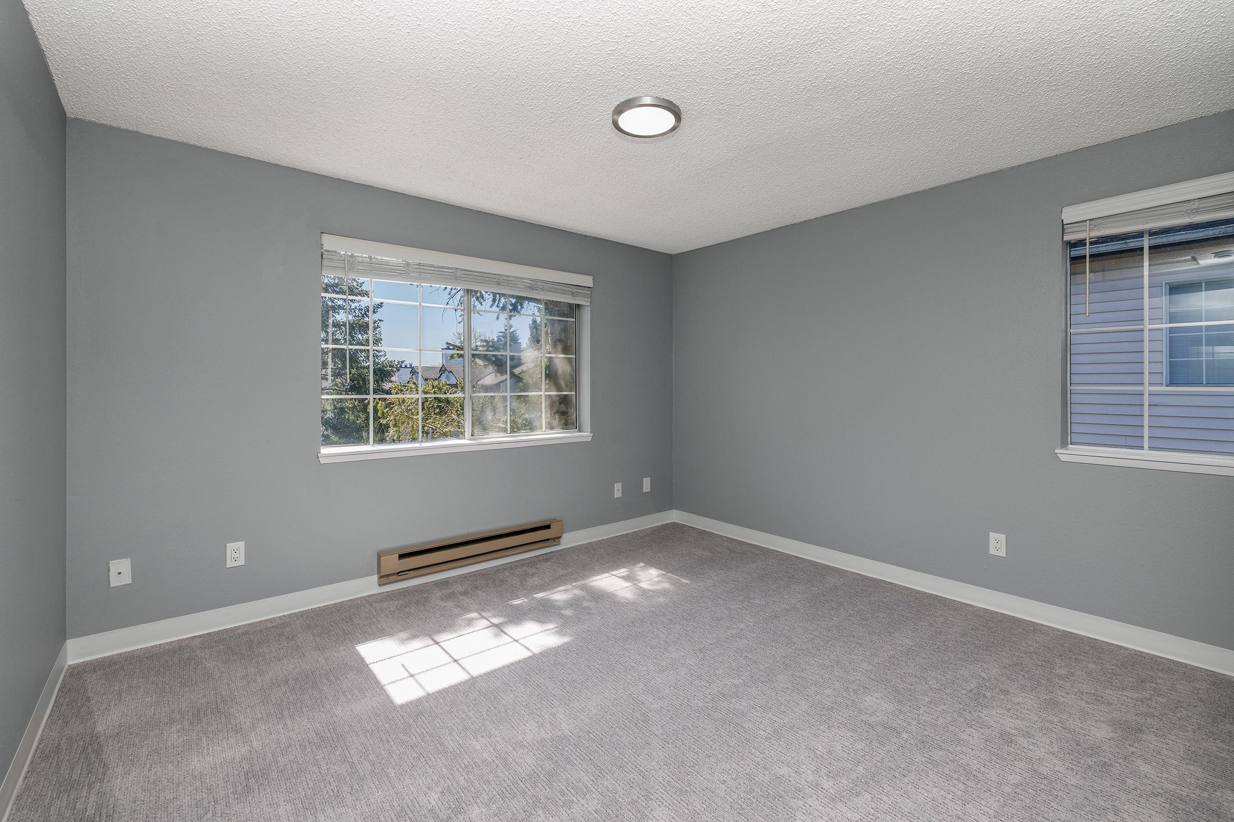 A well-lit, empty room featuring grey walls and carpet. It has two windows that allow natural light to enter, with a ceiling light fixture visible. The space is uncluttered and ready for furniture or decoration.