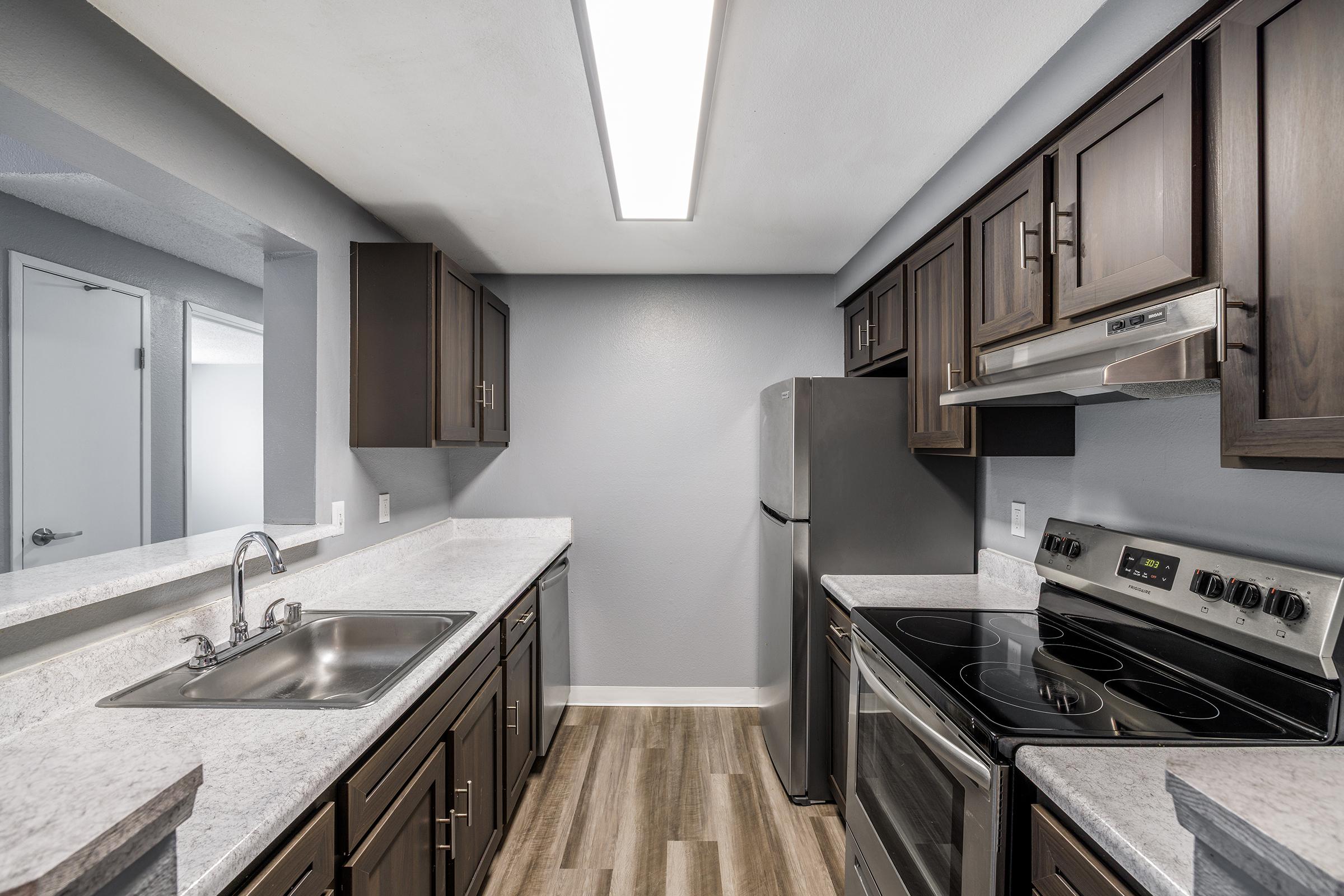 A modern kitchen featuring dark wood cabinets, a stainless steel refrigerator, and an electric stove. The counters are a light-colored stone, and the walls are painted in a soft gray. A long overhead light illuminates the space, creating a bright and functional cooking area.