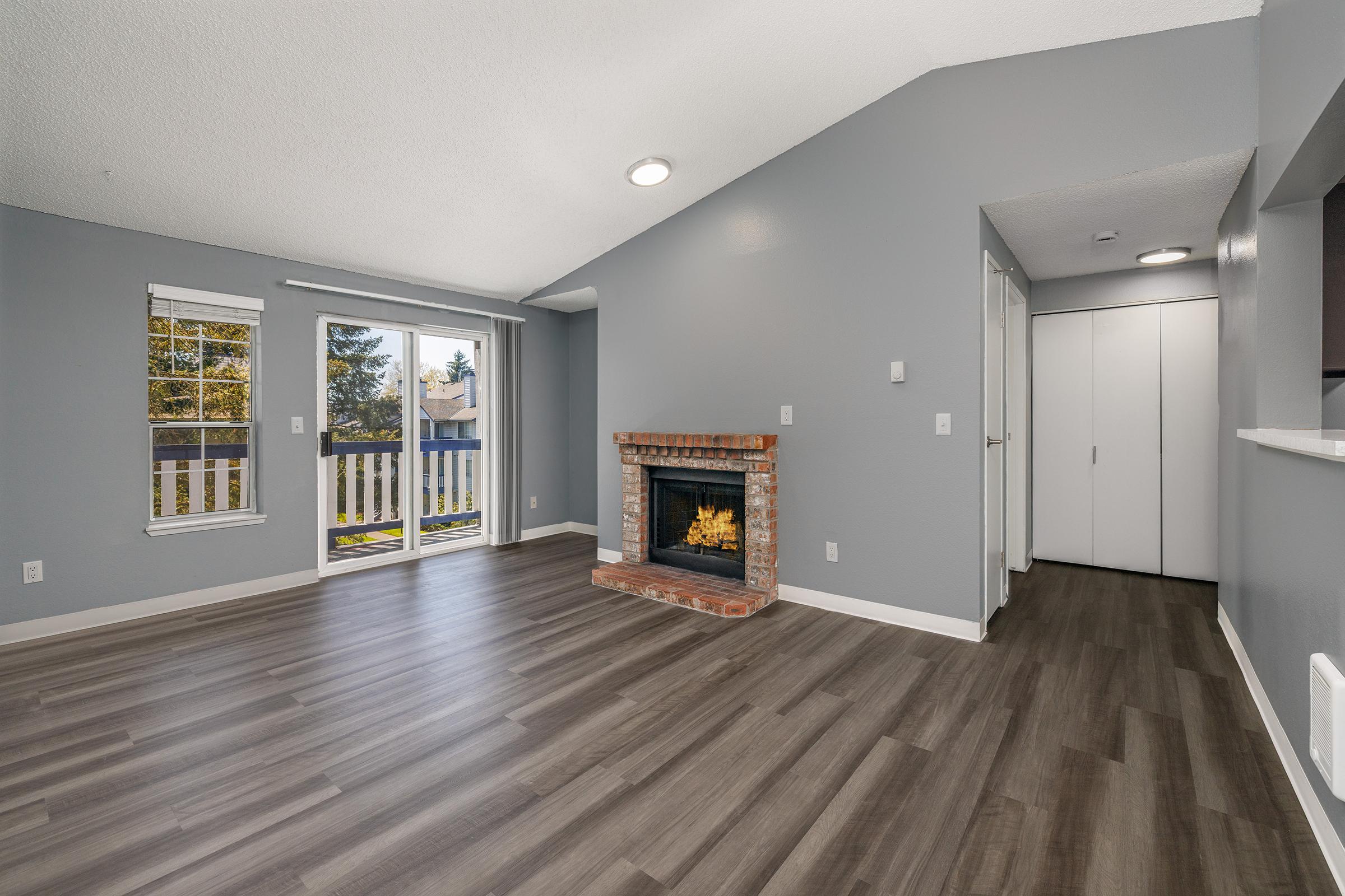 A spacious, modern living room featuring a brick fireplace, large windows with natural light, and laminate flooring. The room is painted in light gray, with a white trim. A sliding glass door leads to a balcony, and there's a hallway with a closet visible on the right.