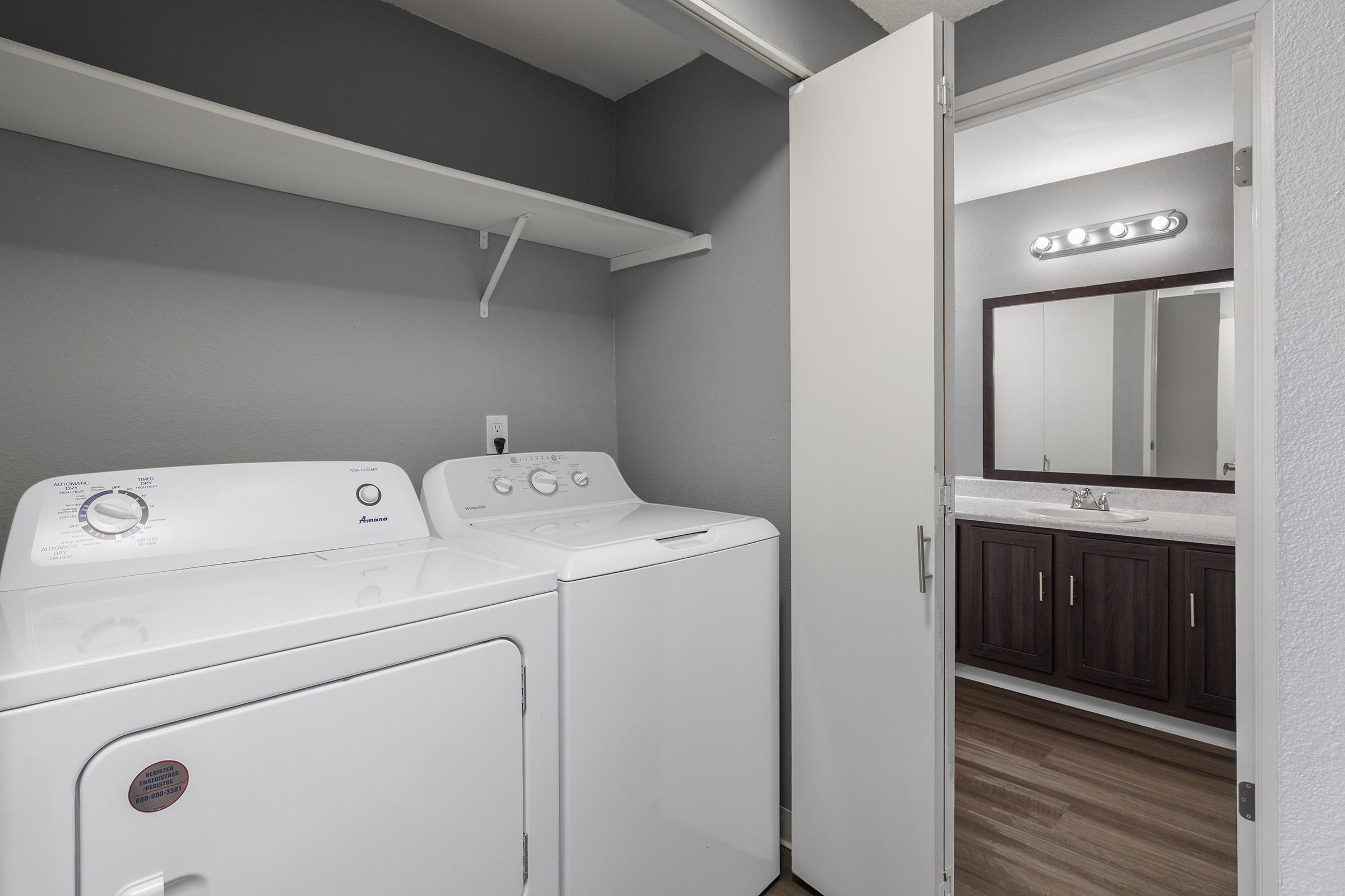 A modern laundry room featuring a white washer and dryer side by side, a shelf above for storage, and an open door leading to a bathroom with a dark wood vanity, sink, and mirror illuminated by overhead lights. The walls are painted gray, creating a clean and contemporary look.