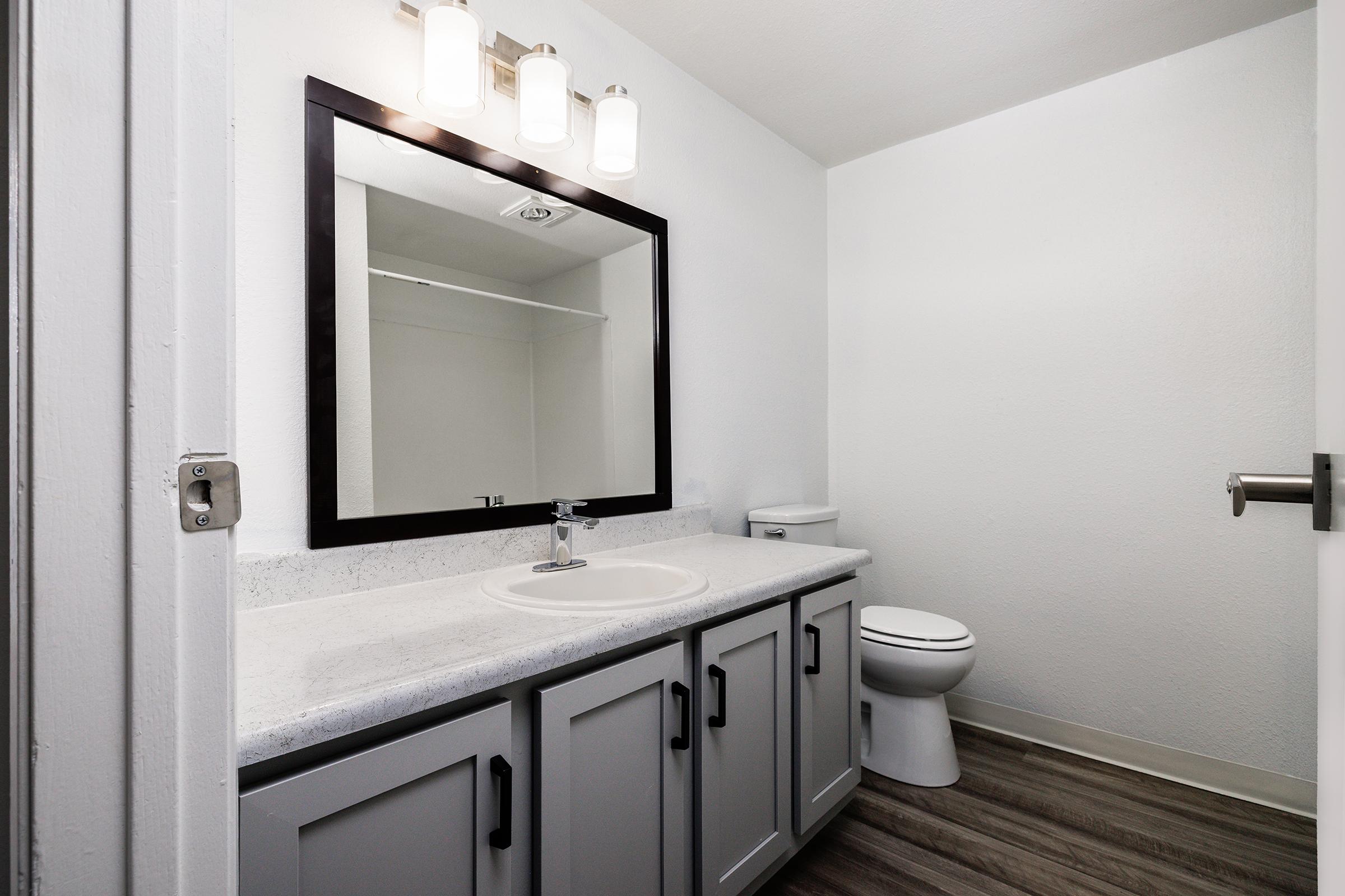 A clean, modern bathroom featuring a dark-framed mirror above a light-colored countertop with a sink. Cabinets are below the countertop, with a white toilet visible to the right. The walls are painted white, and the floor has a wood-like finish. Warm lighting fixtures are mounted above the mirror.