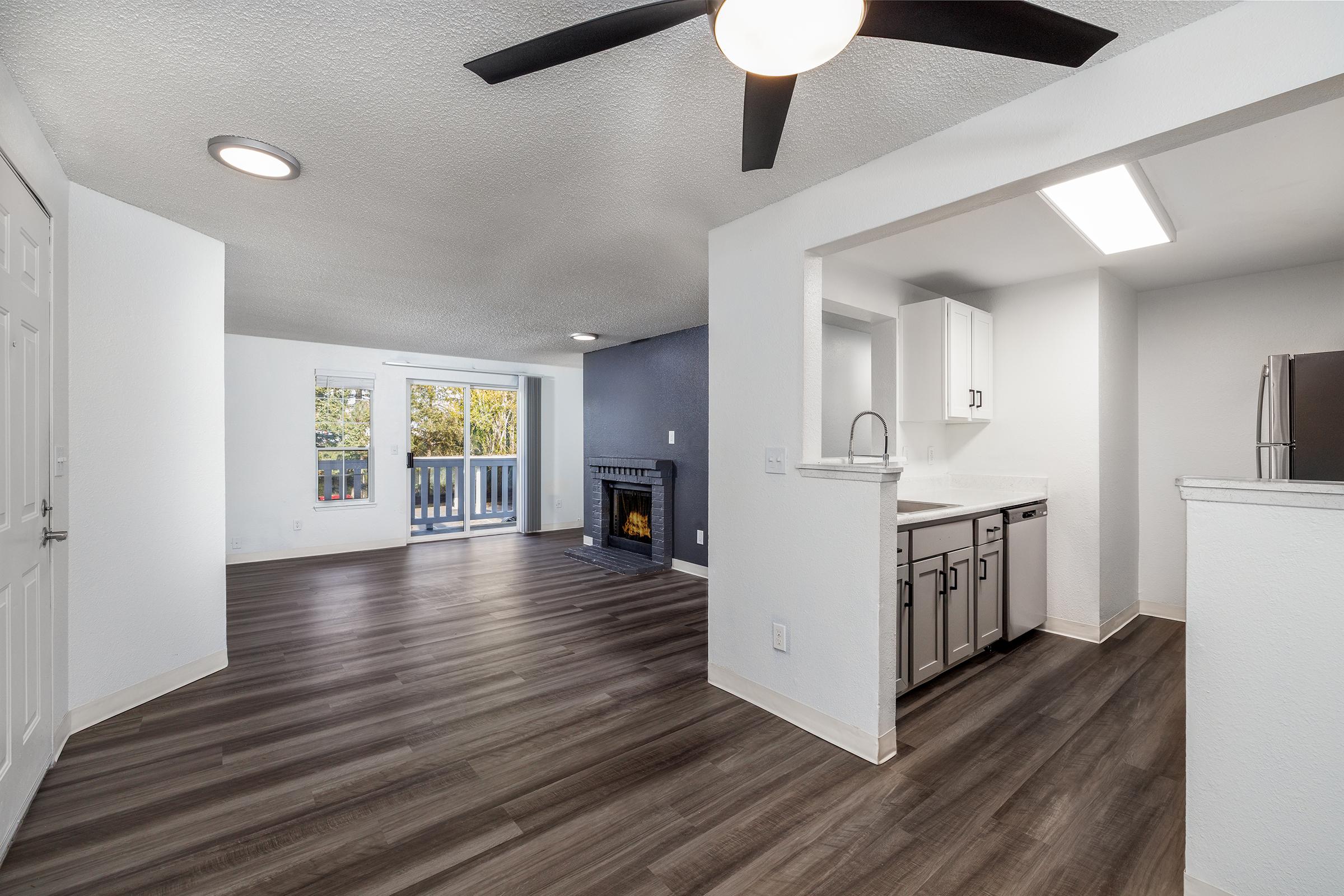 Bright and modern open-concept living space featuring a living room with sliding glass doors, a balcony, and a cozy fireplace. The kitchen is equipped with contemporary cabinets and appliances, with a view of the living area. The flooring is stylish, and the walls are freshly painted in neutral tones.
