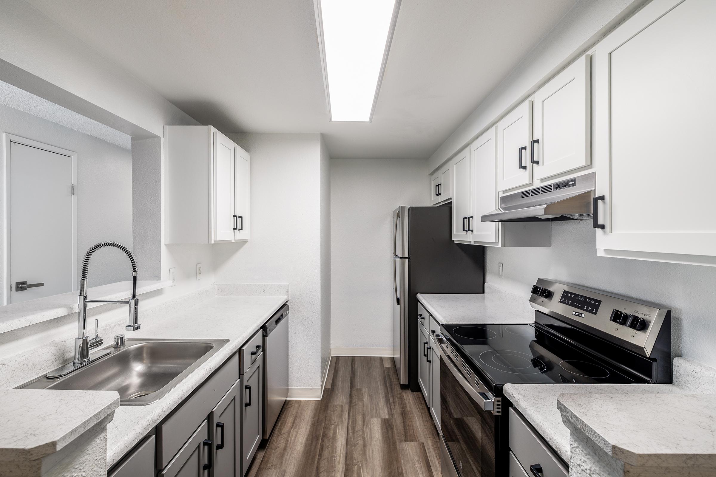 Modern kitchen featuring white cabinets, stainless steel appliances, a double-basin sink, and a sleek countertop. The space is well-lit with a long overhead light, showcasing a clean and minimalist design.