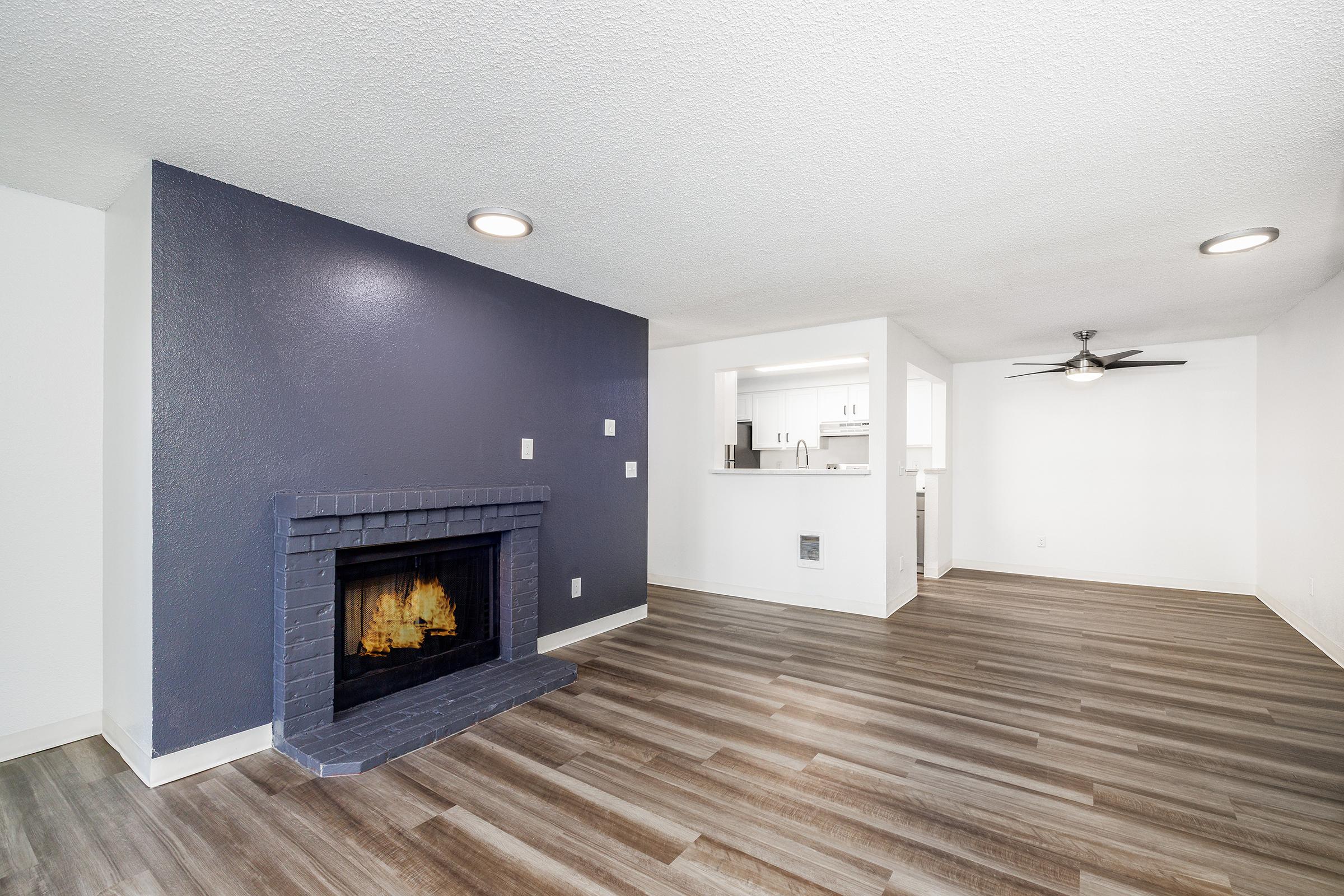 A modern living room featuring a dark accent wall with a fireplace, light-colored walls, and laminate flooring. The space is open to a kitchen area visible in the background, which includes white cabinetry. A ceiling fan is present, providing a bright and airy atmosphere.