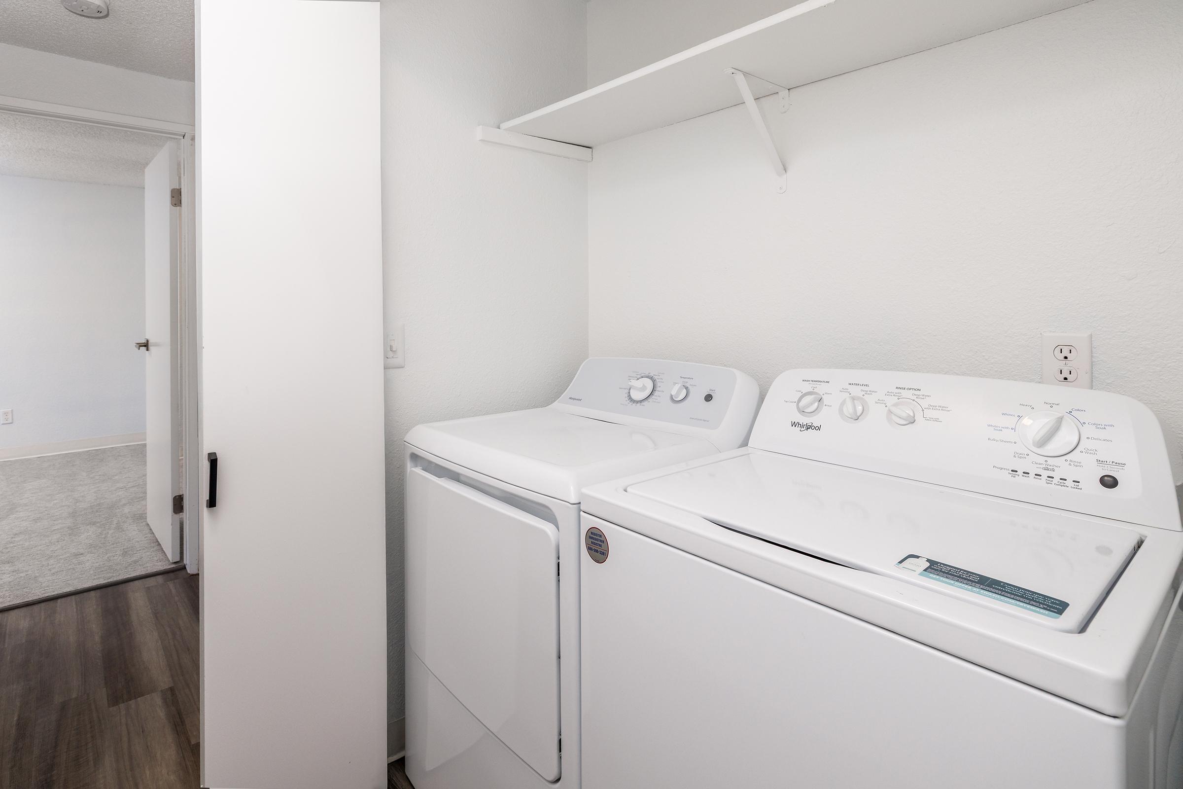 A laundry area featuring a white washer and dryer side by side, with a shelf above for storage. The walls are painted light gray, and there is a doorway leading to another room in the background. The floor has a wood-like finish, and the space appears clean and organized.