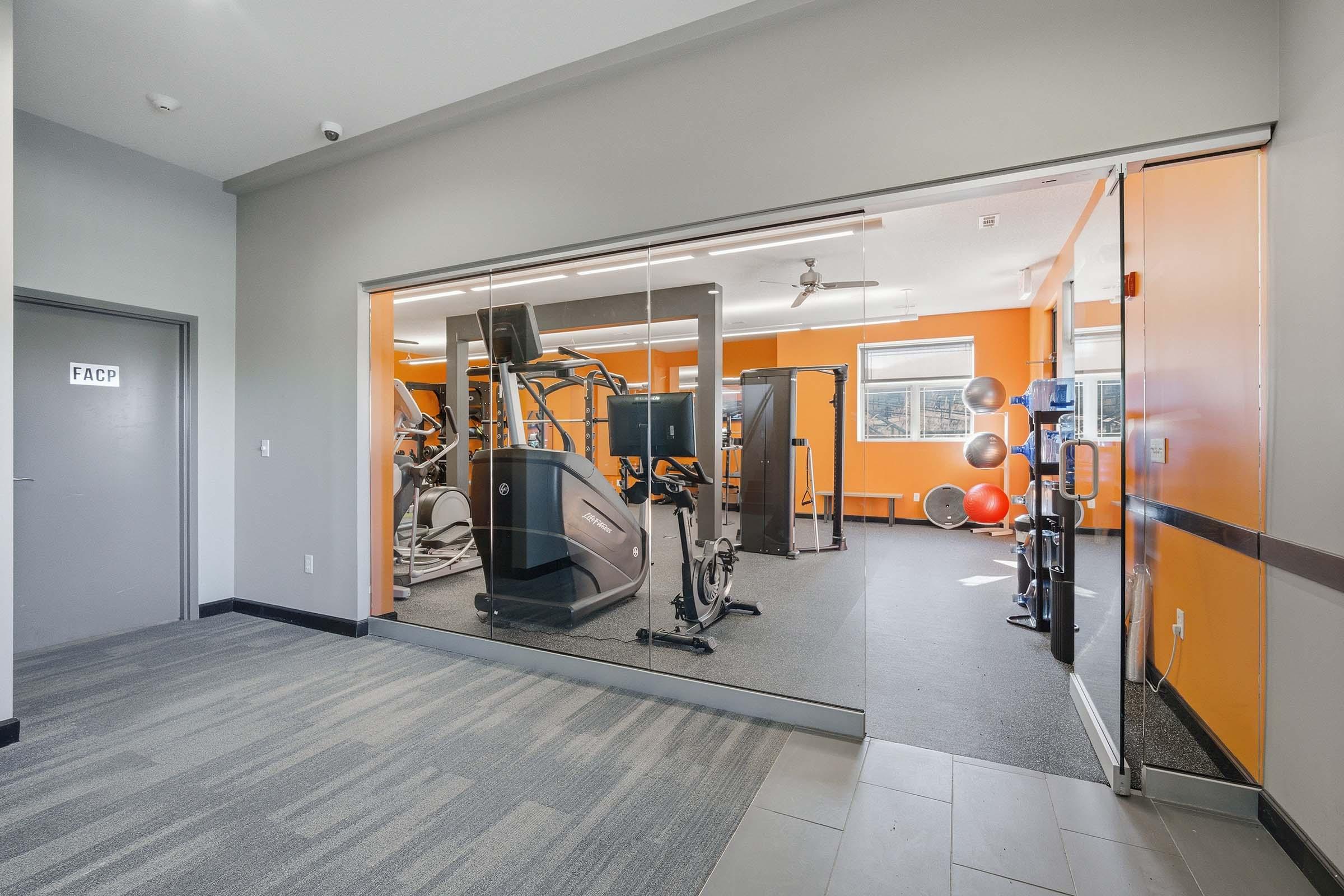 A view into a modern gym featuring exercise equipment such as treadmills, stationary bikes, and exercise balls. The walls are painted orange, and there is a large glass window displaying the workout area. The flooring is black and gray, creating a contemporary atmosphere.