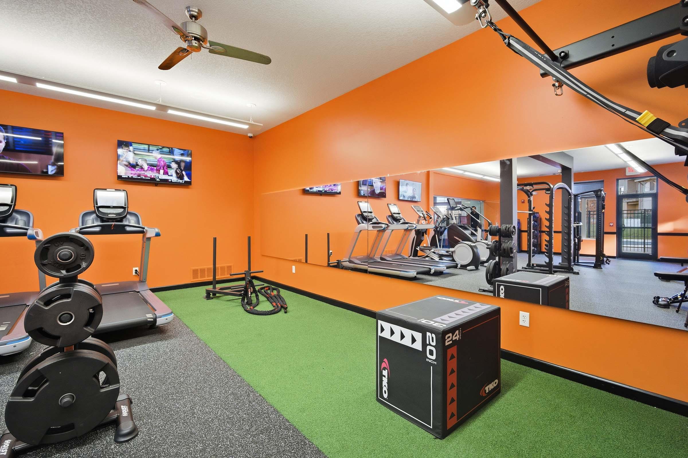 Modern gym interior featuring cardio equipment like treadmills and a mirror along one wall. The bright orange walls enhance the lively atmosphere, while a variety of fitness accessories, including weights and a workout box, are visible on the green turf floor. Two TVs are mounted on the opposite wall.