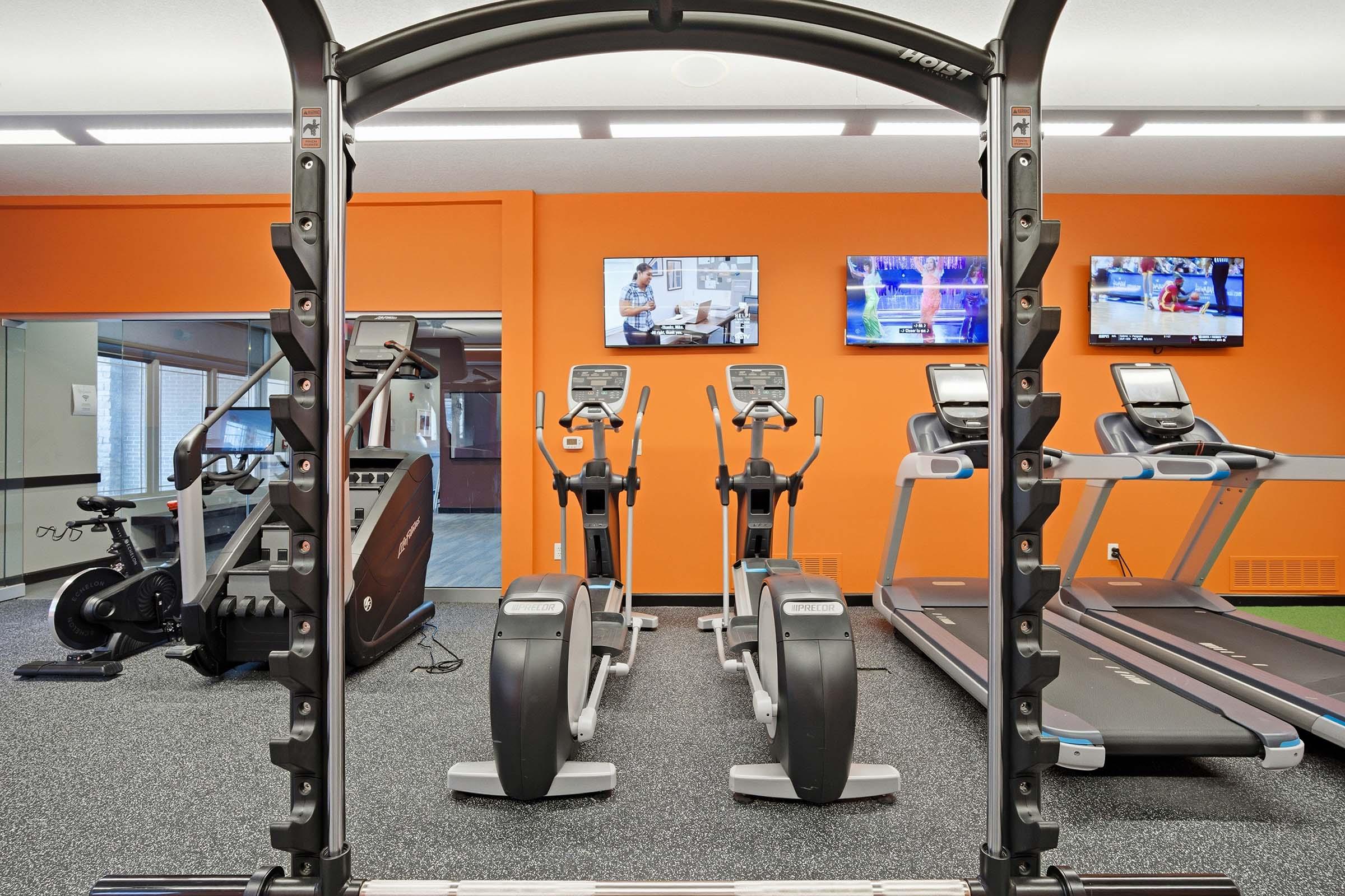A modern gym interior featuring a squat rack in the foreground. In the background, there are two treadmills and two elliptical machines, all set against an orange wall. Two televisions are mounted on the wall, displaying workout videos. The flooring is black with a textured finish.