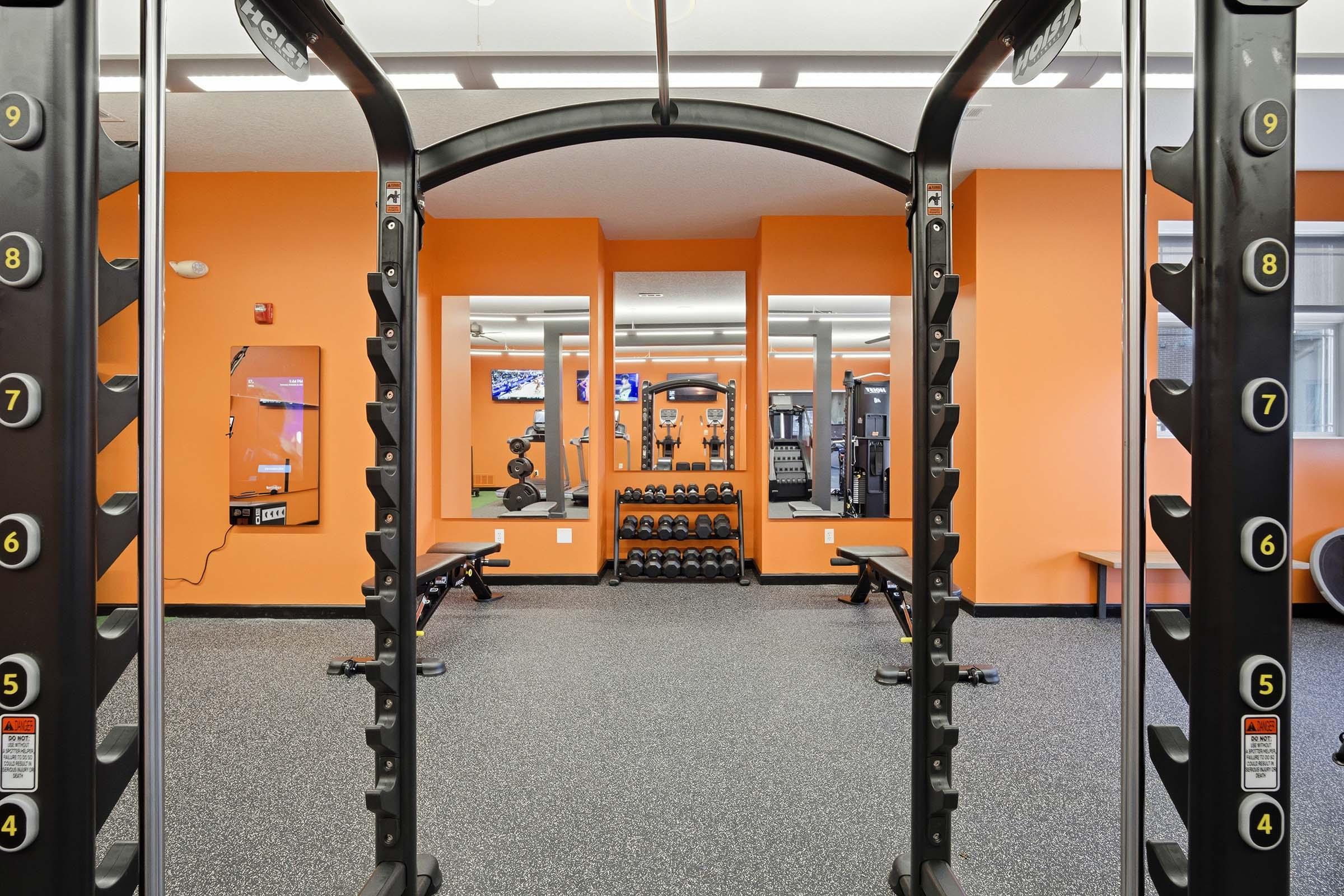 A modern gym interior featuring a weight rack with dumbbells, benches, and various strength training equipment. The walls are painted orange, and there is ample natural light. The floor is covered in black rubber flooring, enhancing the workout environment.