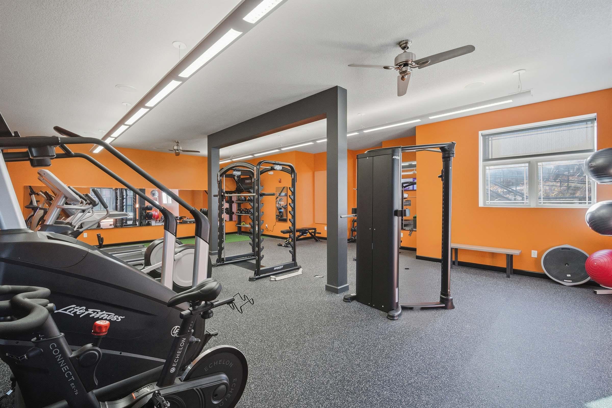 A modern gym interior featuring exercise equipment such as a stationary bike, strength training machines, and free weights. The walls are painted orange, and there are mirrors and floor mats visible, creating a bright and inviting workout space.