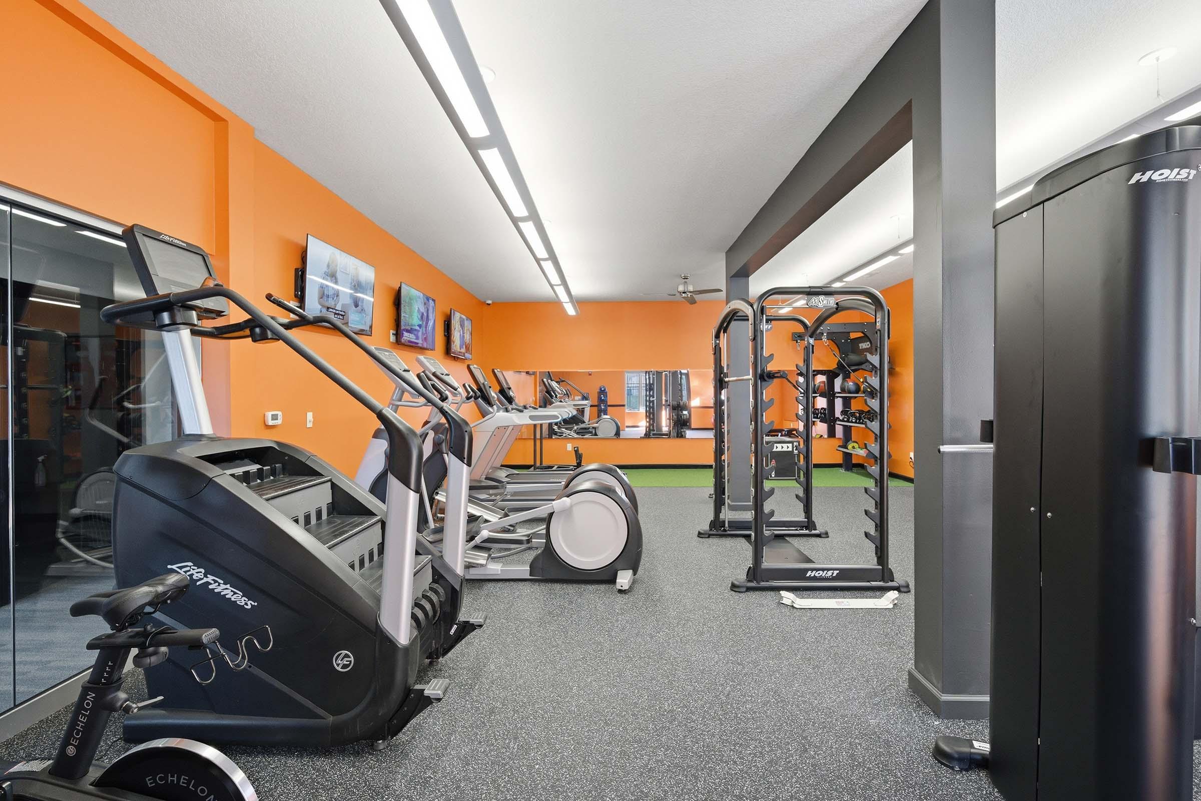 A modern gym interior featuring exercise equipment including treadmills, an elliptical machine, a set of weights on a rack, and a water cooler. The walls are painted orange, and there are televisions mounted for entertainment. The flooring is dark with sections of green turf.